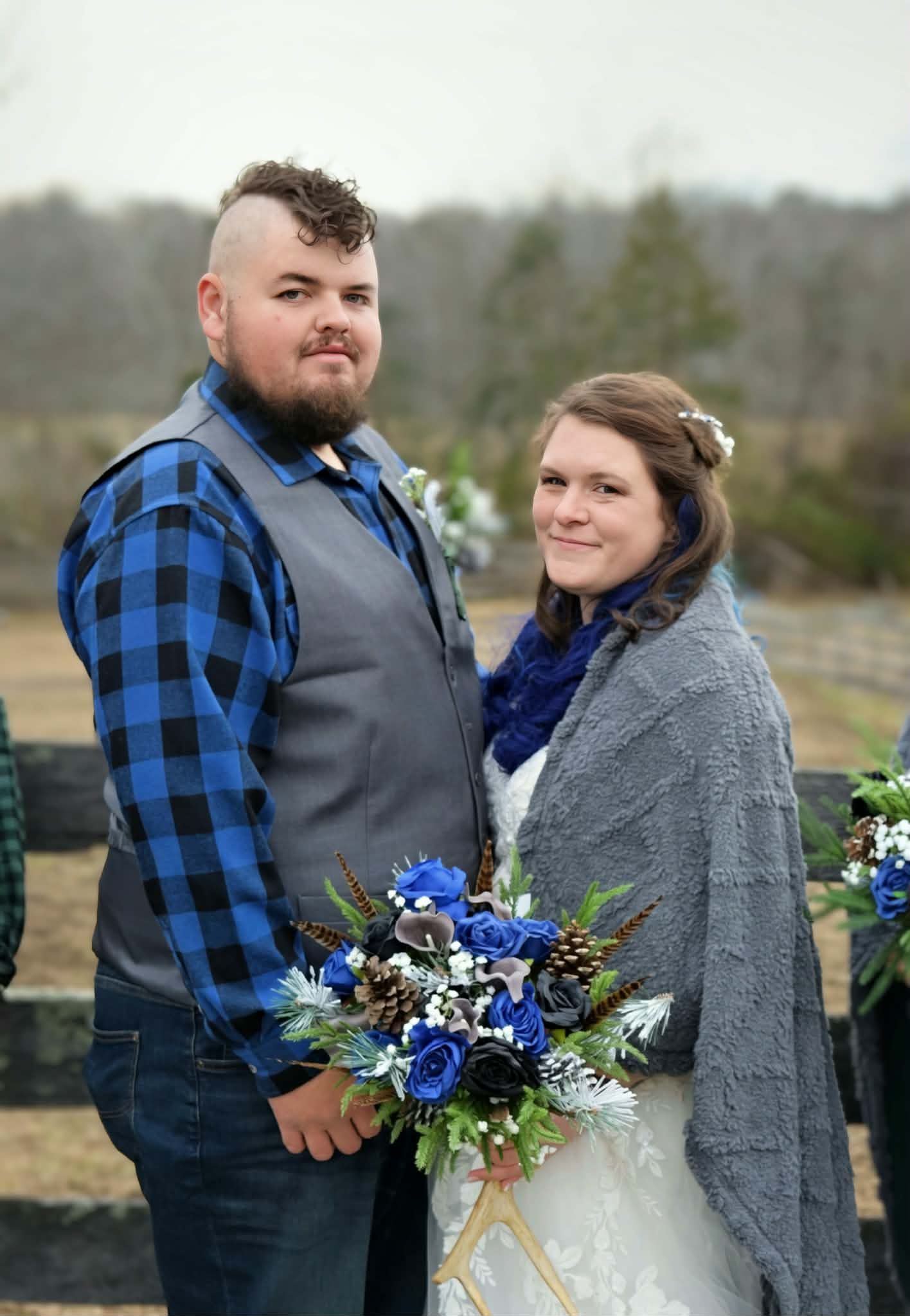 Bride and groom posing outdoors at winter wedding, groom in plaid shirt and vest, bride holding blue floral bouquet.