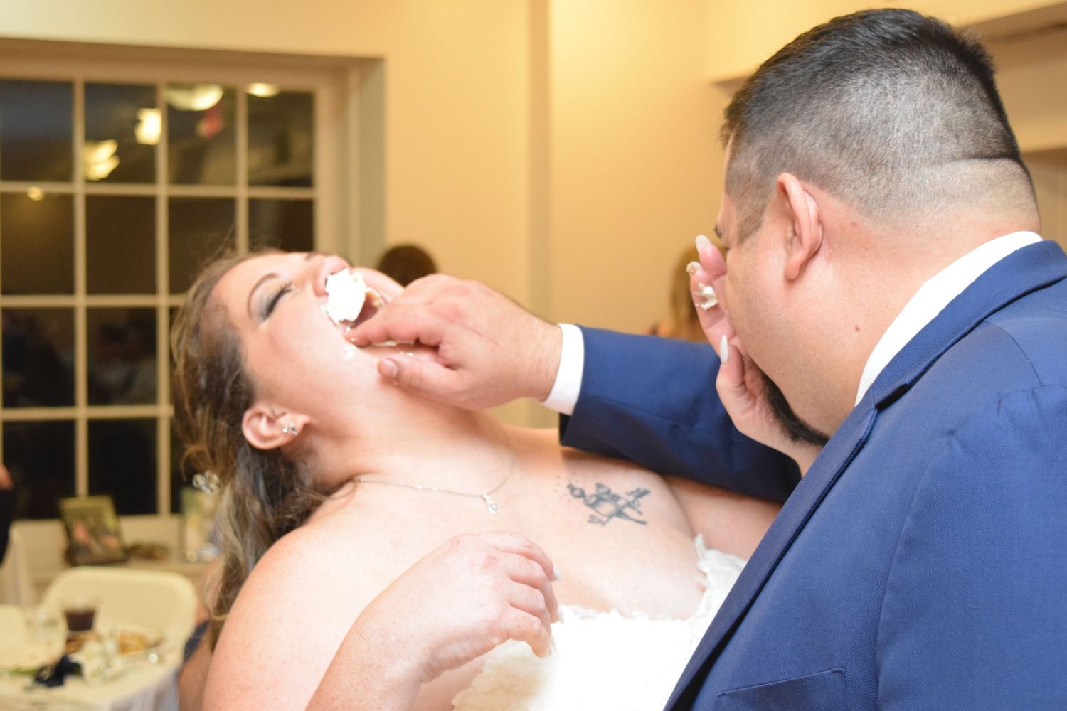 Groom feeding bride a bite of wedding cake during reception, both smiling and wearing formal wedding attire.