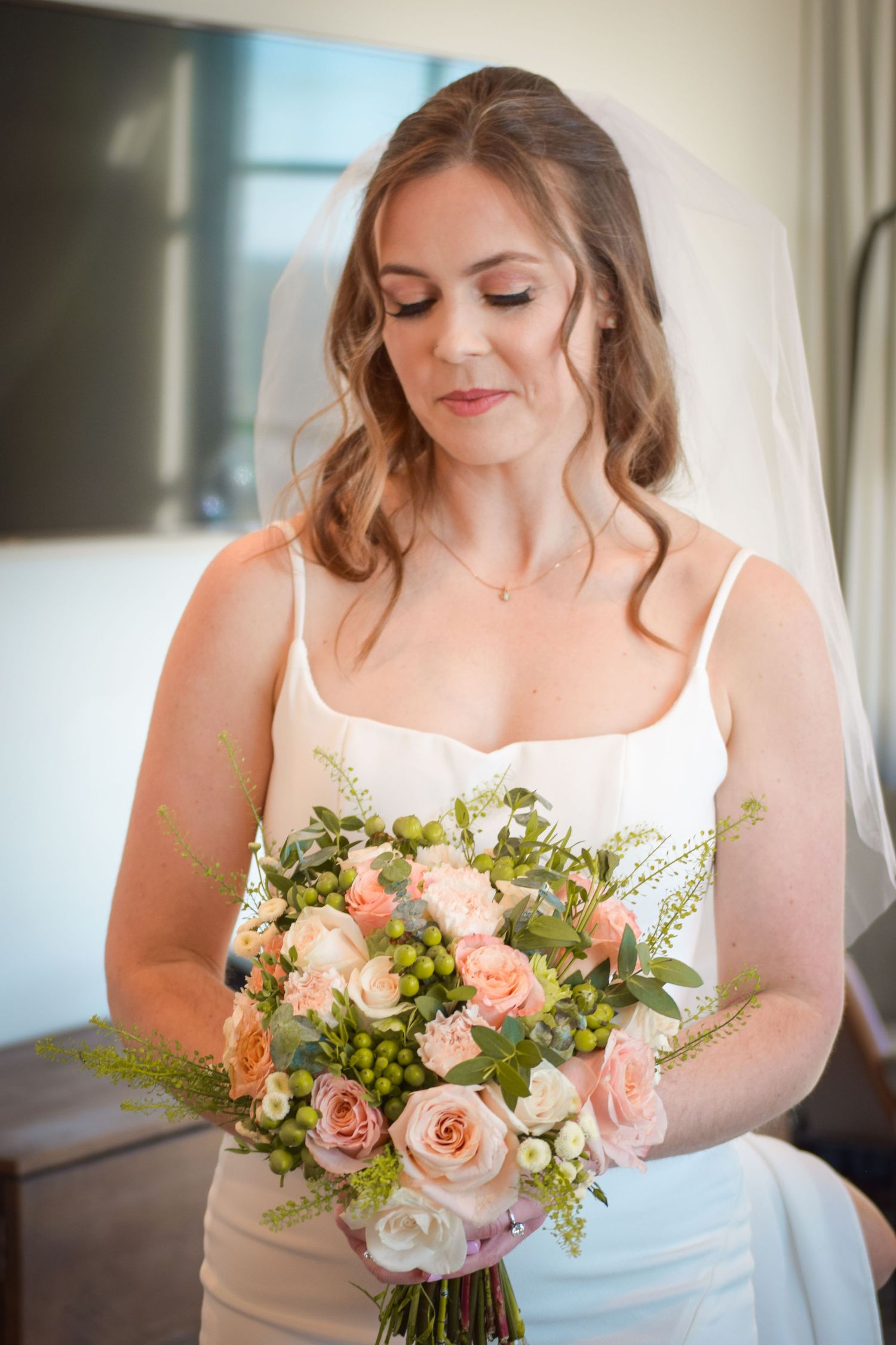 Bride in white veil looking down at peach rose and greenery bridal bouquet.