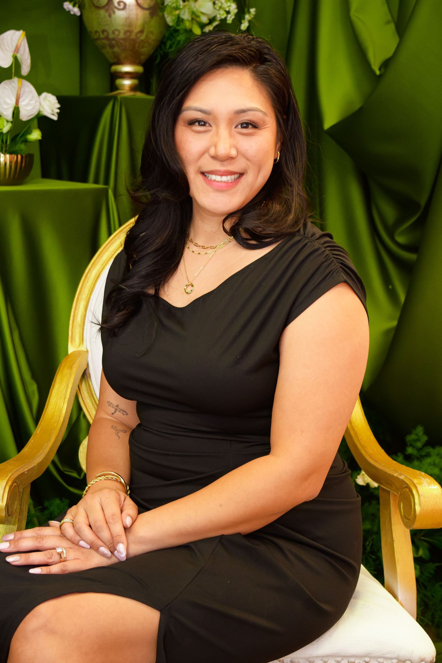 Woman in black dress smiling seated on gold chair with green draped backdrop and white flowers.