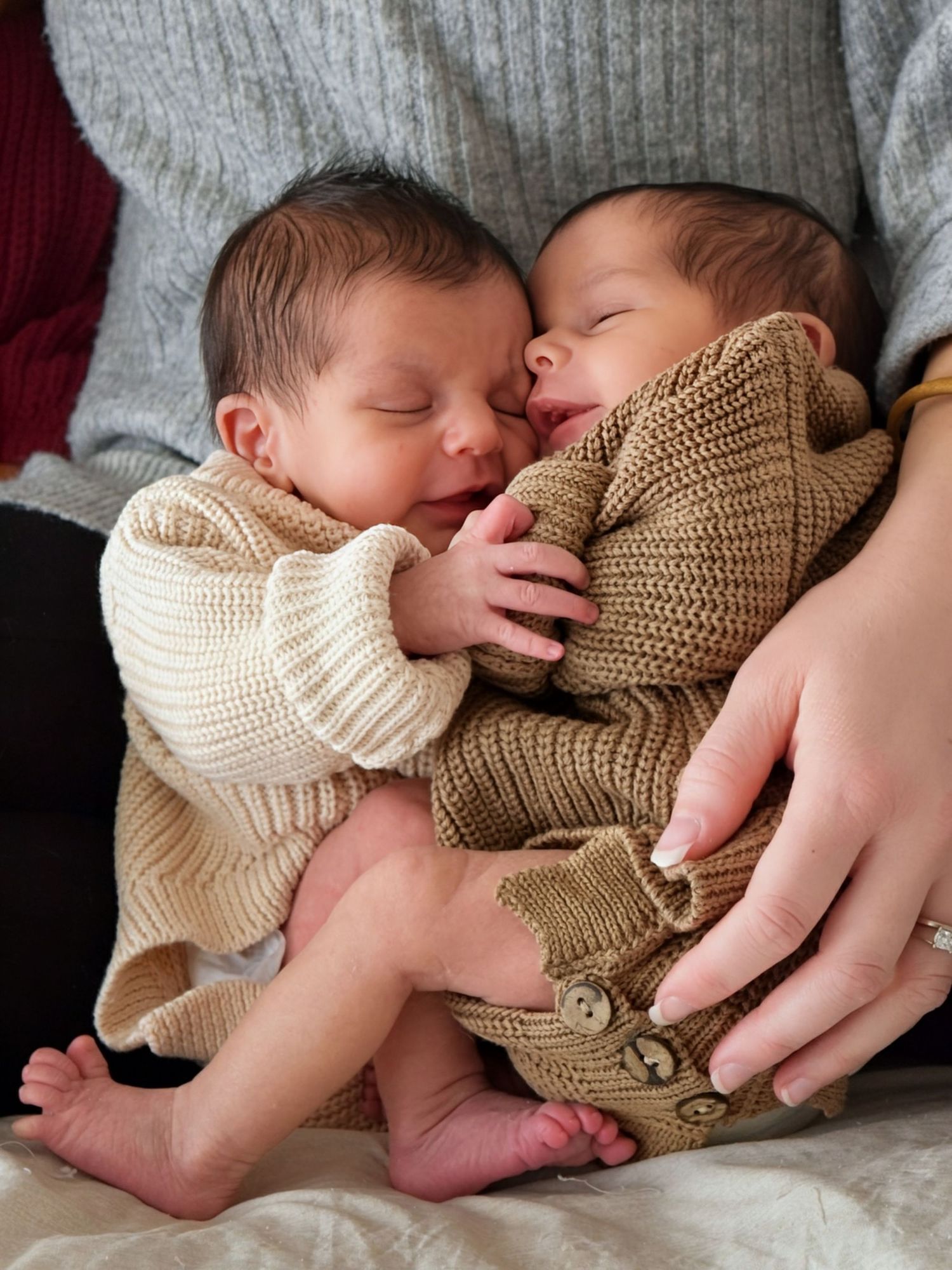 Two sleeping newborn twins in cream and brown knit sweaters cuddled together in parents hands.