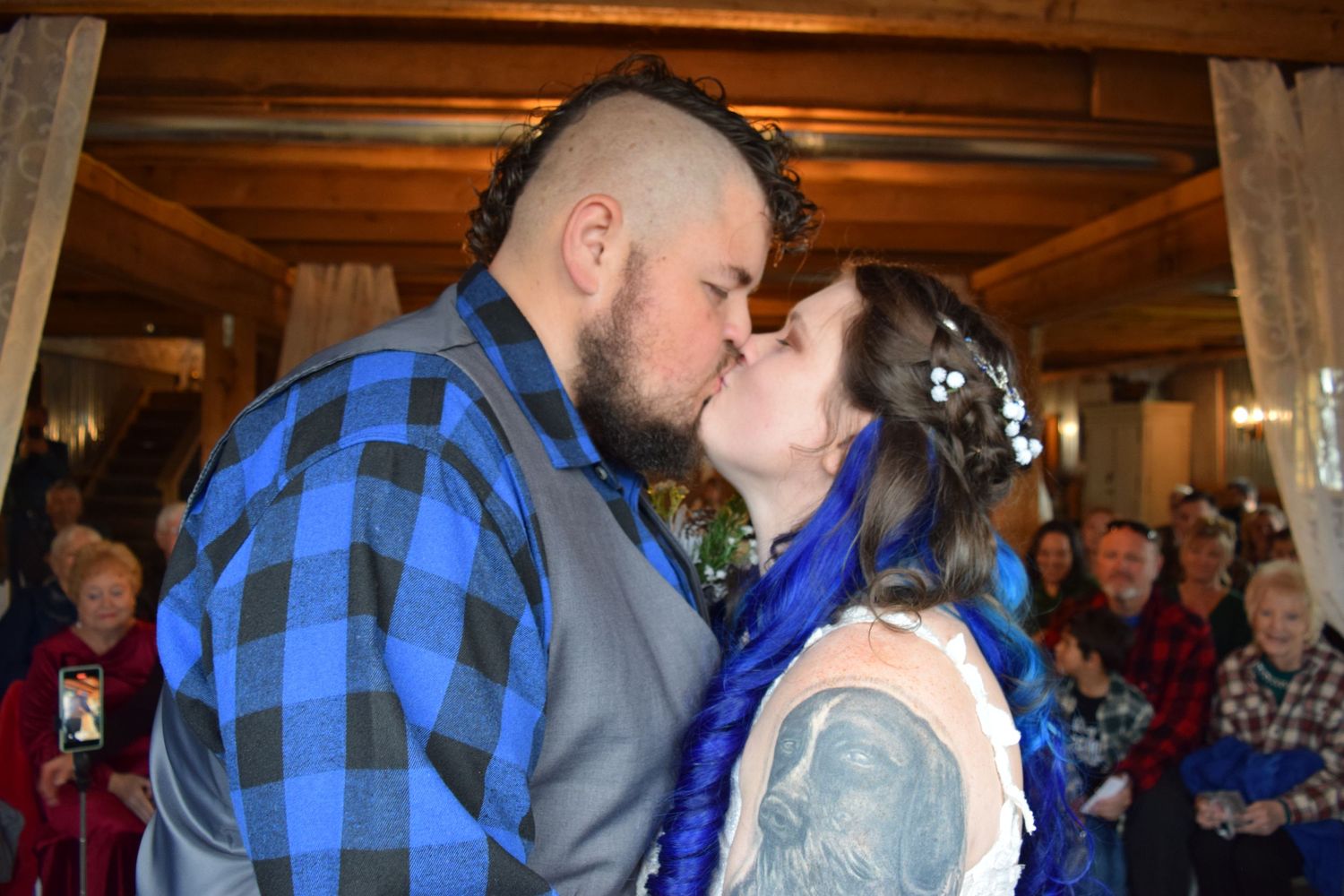 Newlyweds sharing first kiss at wedding ceremony with guests applauding in background.
