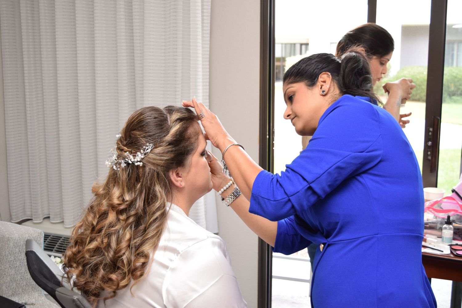 Bridal stylist in blue dress adjusting bride's hair accessories during wedding preparation.