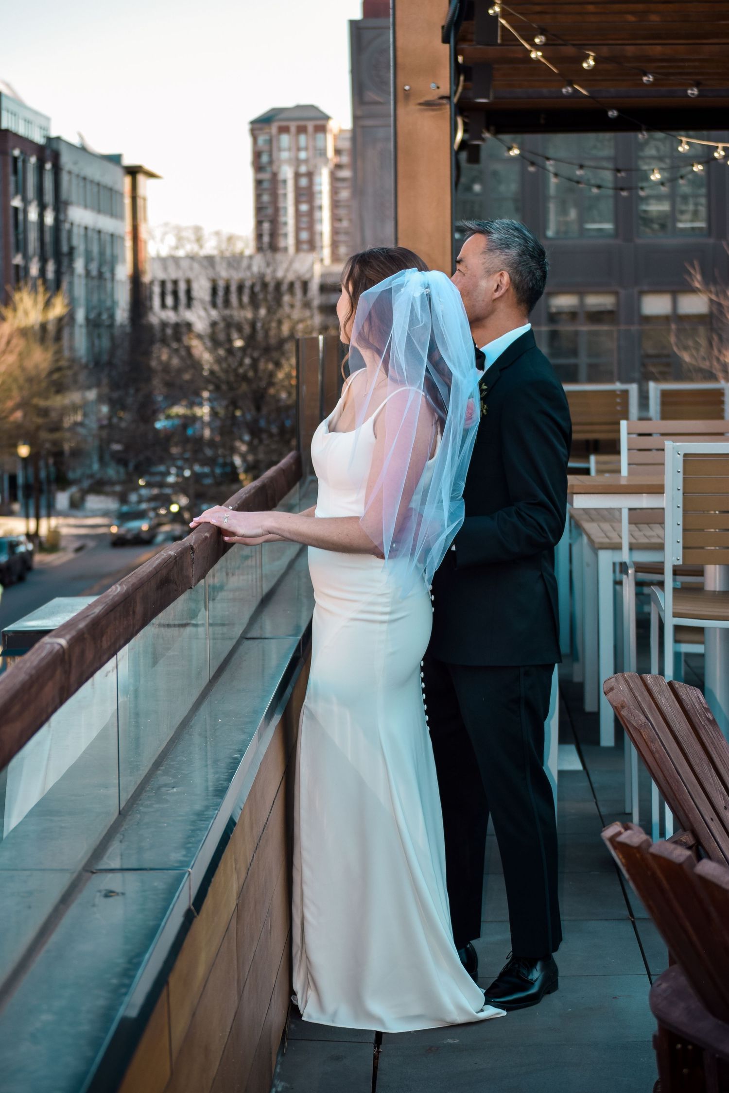 Groom in black tuxedo kisses bride in white gown on rooftop balcony overlooking city street.