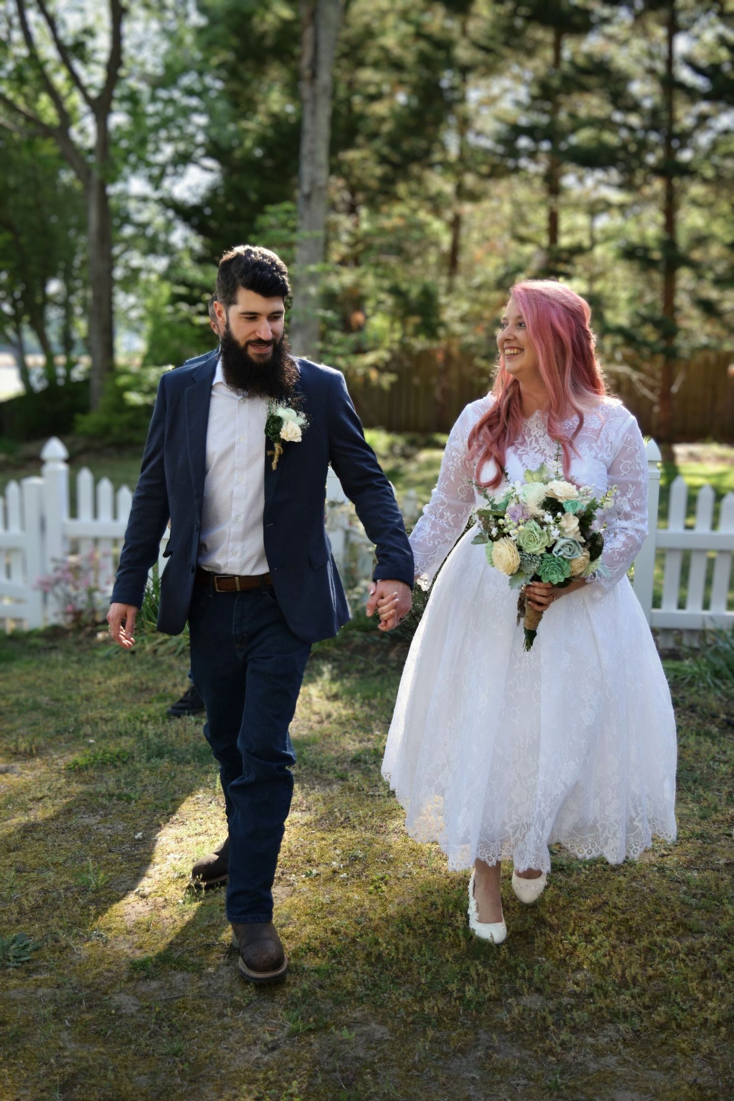 Newlywed couple walking hand in hand outdoors, bride in lace dress with pink hair holding bouquet, groom in navy suit.