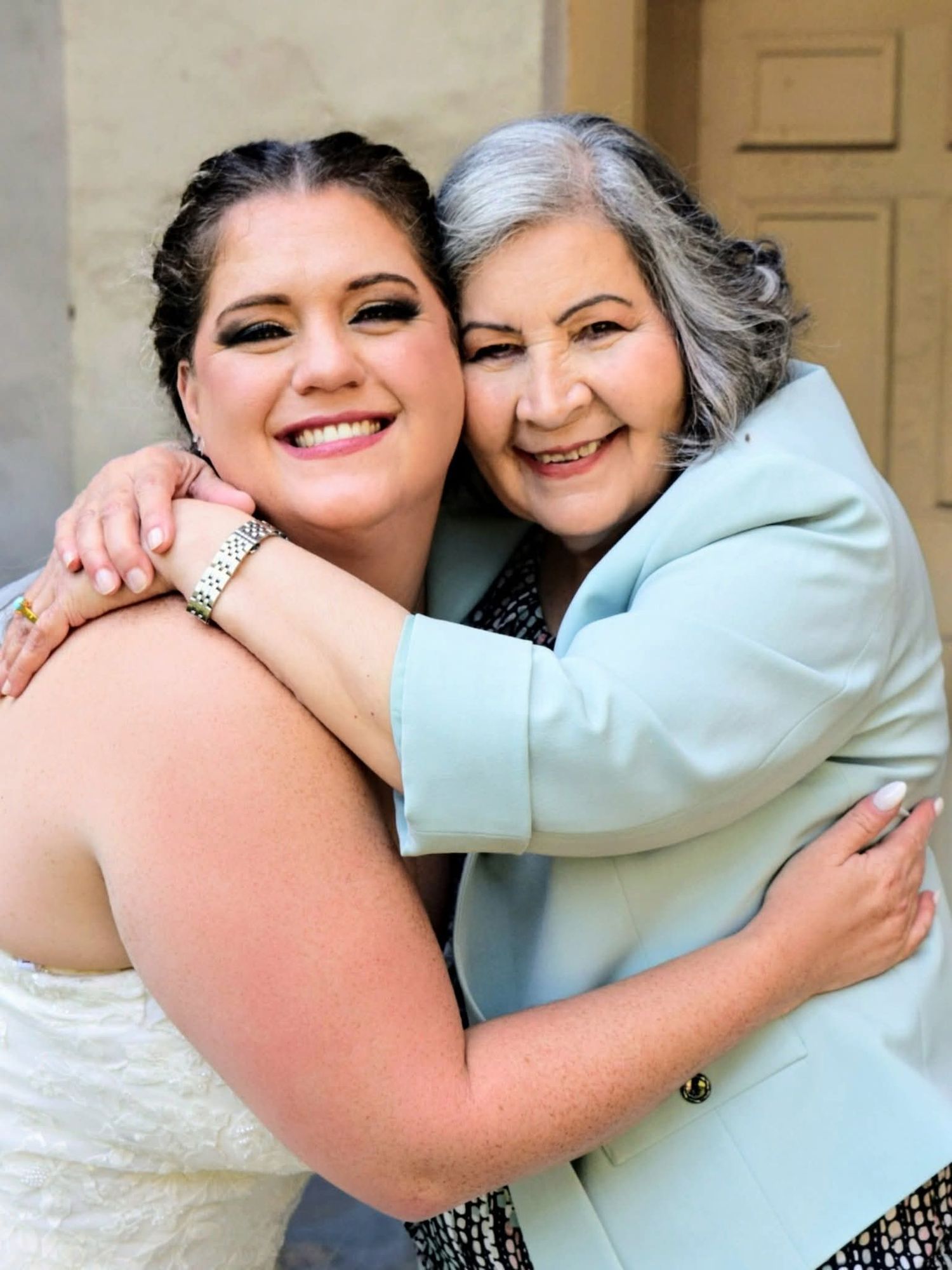 Smiling bride in white gown embraces an older woman in a light blue jacket at wedding celebration.