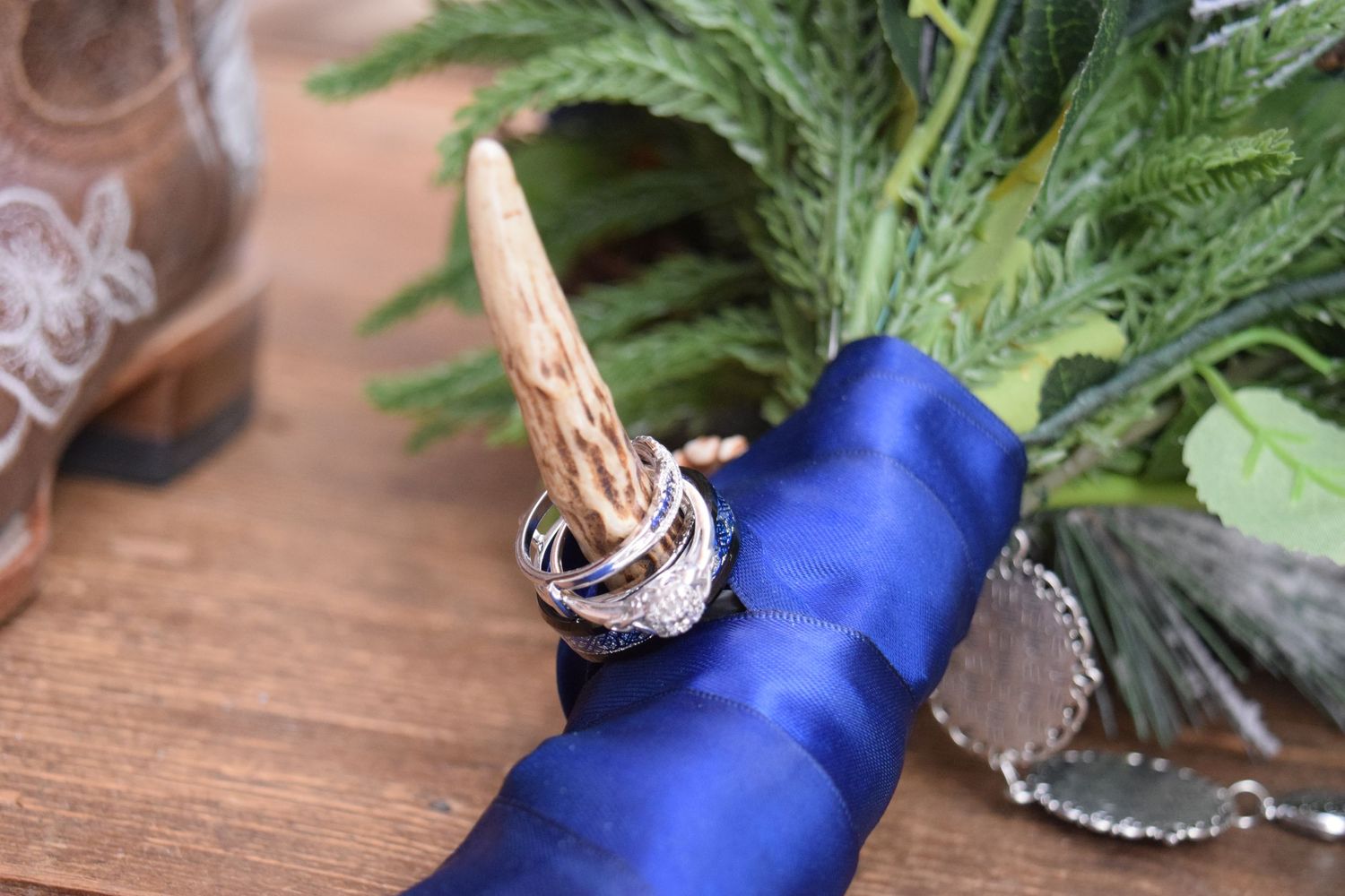 Wedding rings displayed on a deer antler wrapped in royal blue satin ribbon beside a green bouquet.