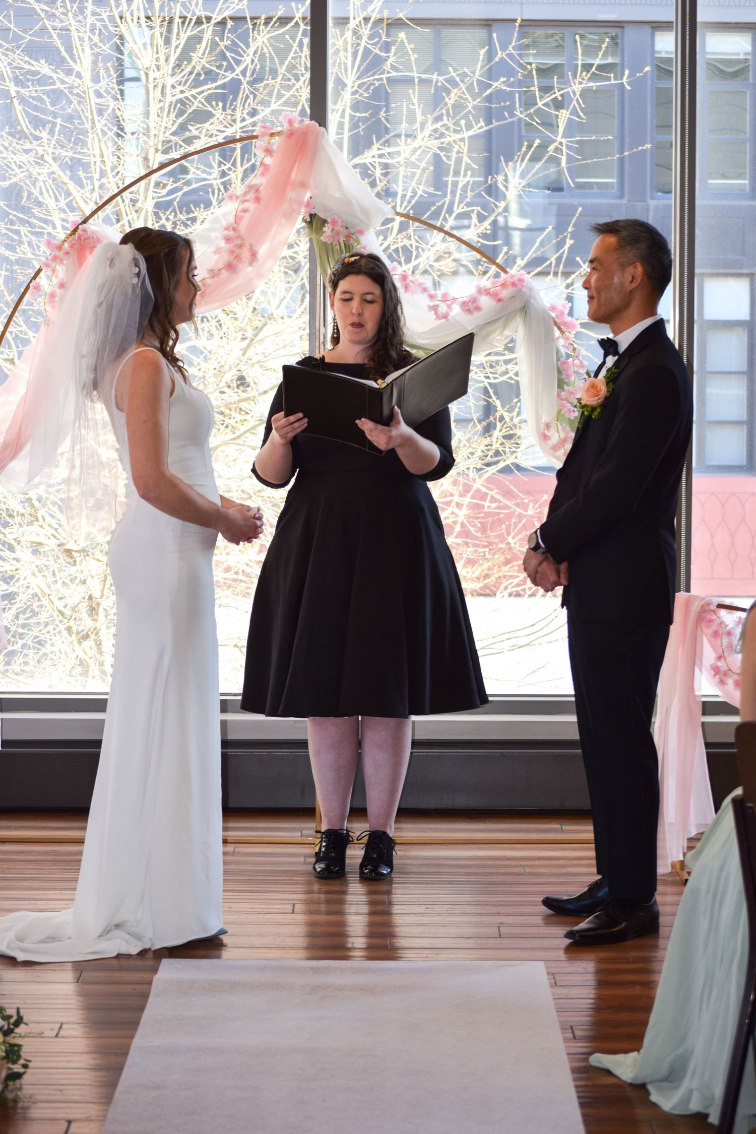 Bride and groom exchange vows before officiant under floral arch at indoor wedding ceremony.
