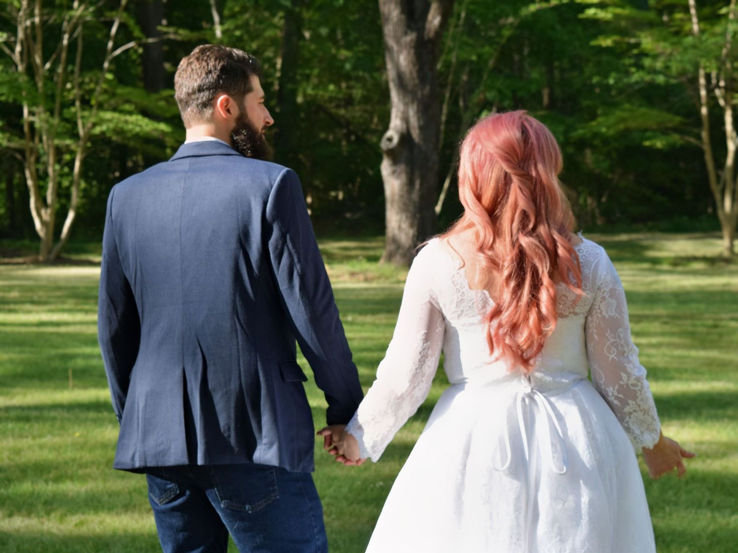 Newlywed couple walking hand in hand through green park, bride in white lace dress with pink hair, groom in navy suit.