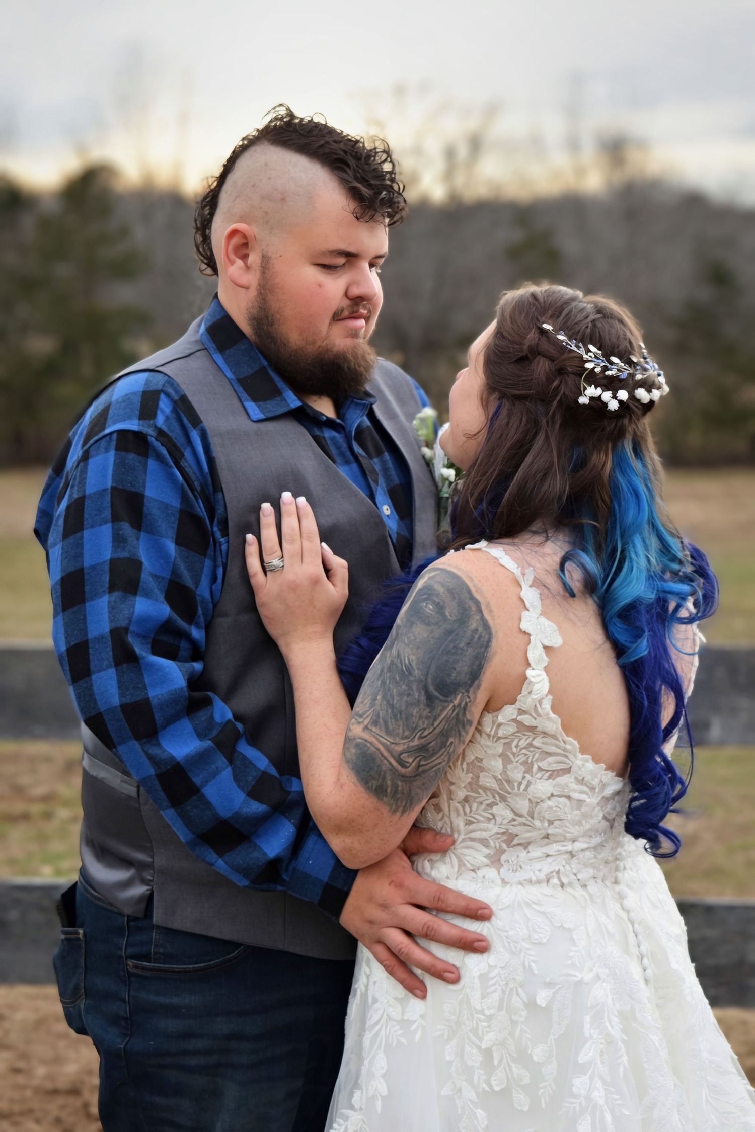 Groom in blue plaid shirt looking lovingly at bride in lace backless gown during outdoor portrait.
