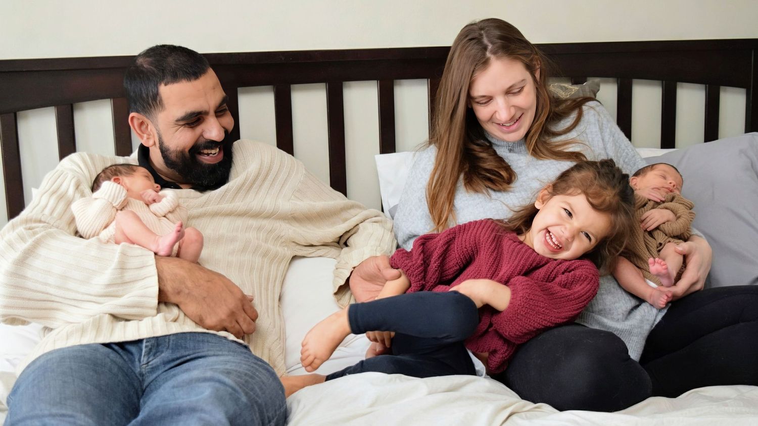 Happy family relaxing on bed with newborn baby and toddler, smiling parents enjoying a cozy moment together.