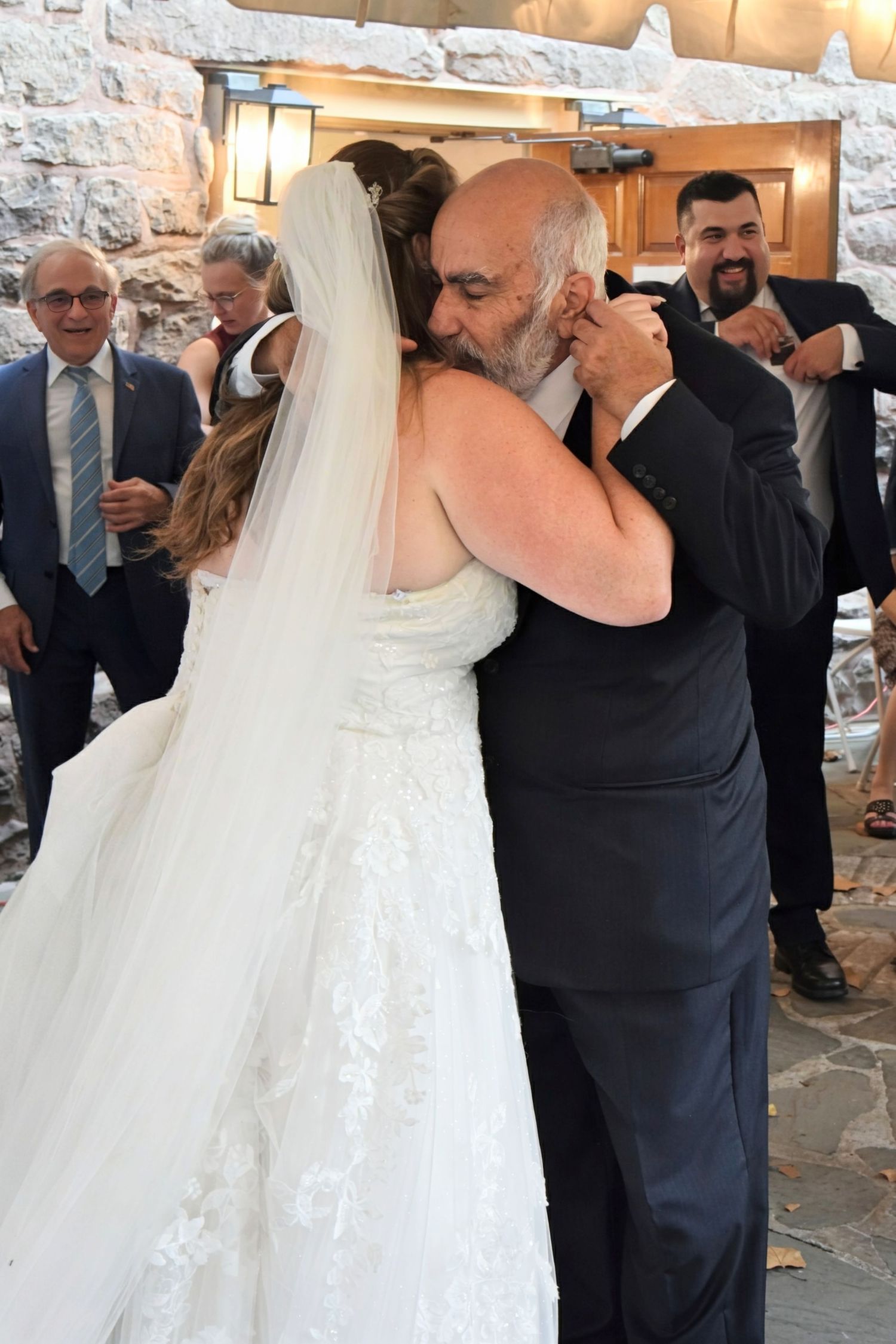 Emotional bride in white lace gown sharing heartfelt hug with older man at wedding reception.
