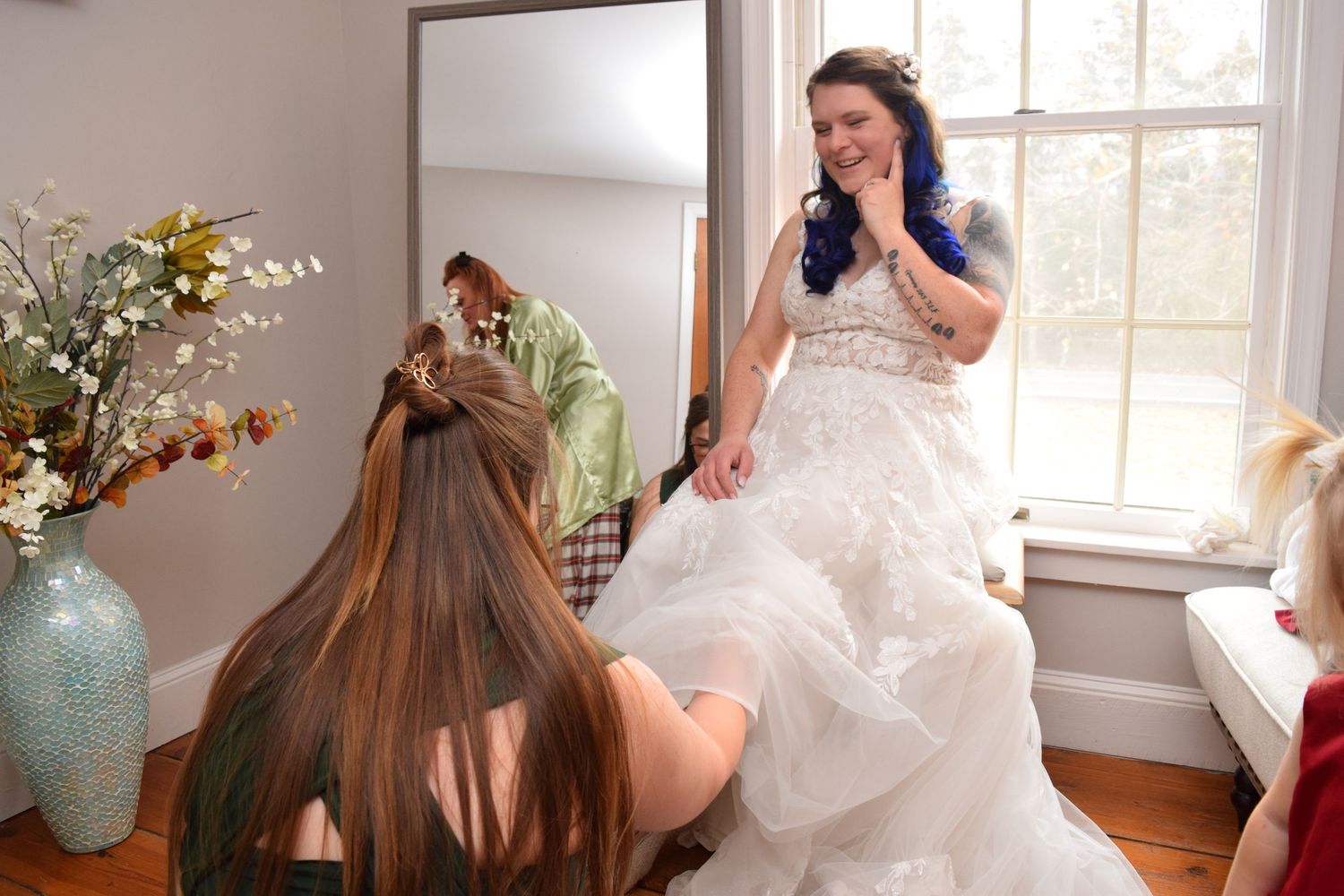 Bridesmaids helping bride in white lace gown with blue hair adjust her dress indoors.