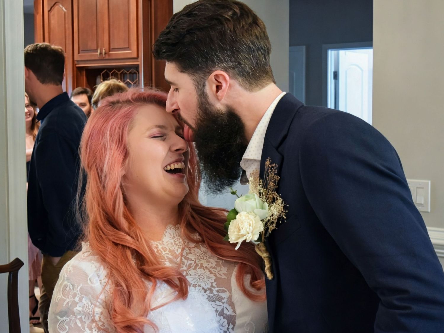 Laughing bride with pink hair and bearded groom sharing a joyful moment together indoors at wedding reception.