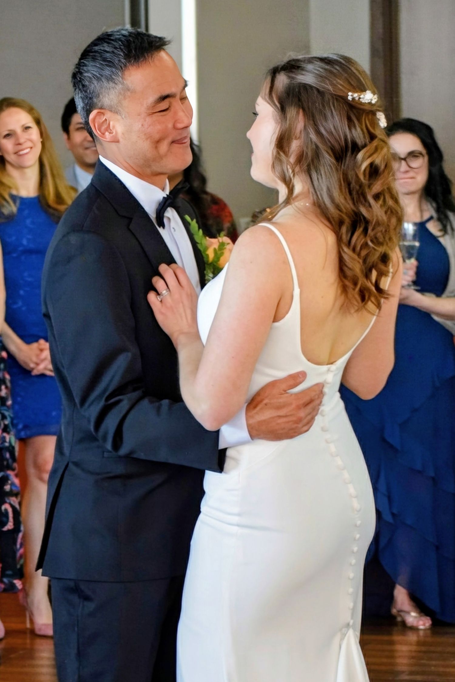 Bride and groom share their first dance, with wedding guests in blue dresses watching in the background.