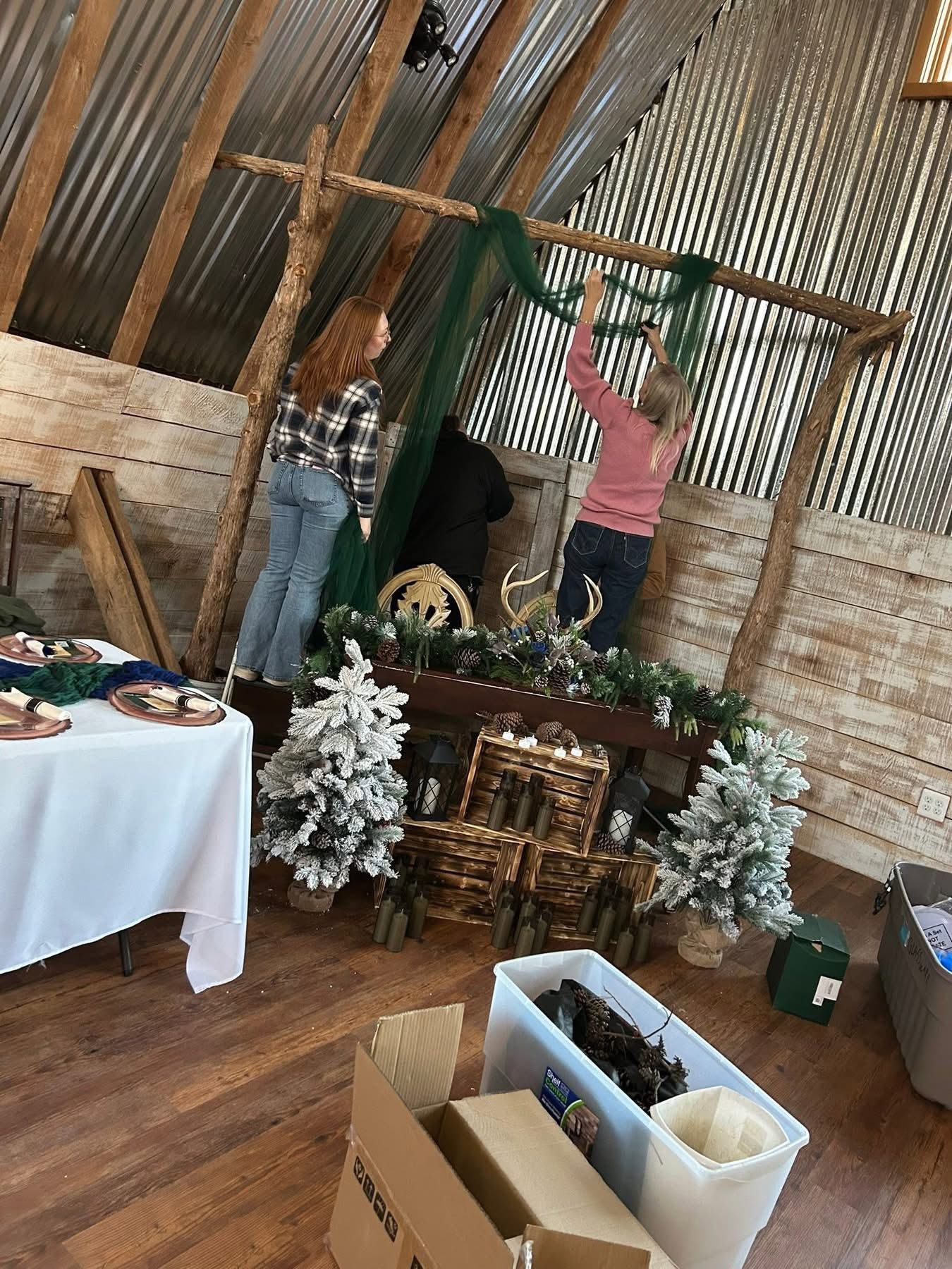 People decorating a rustic barn venue with flocked trees, wooden crates, and green fabric arch.