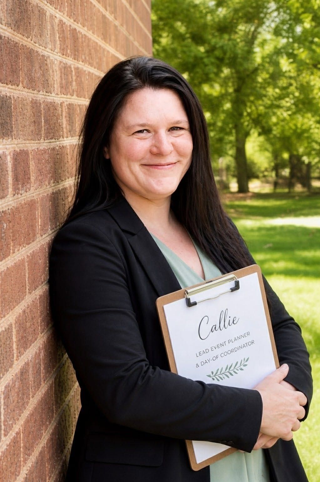 Professional event planner Callie smiles holding a clipboard while leaning against a brick wall outdoors.