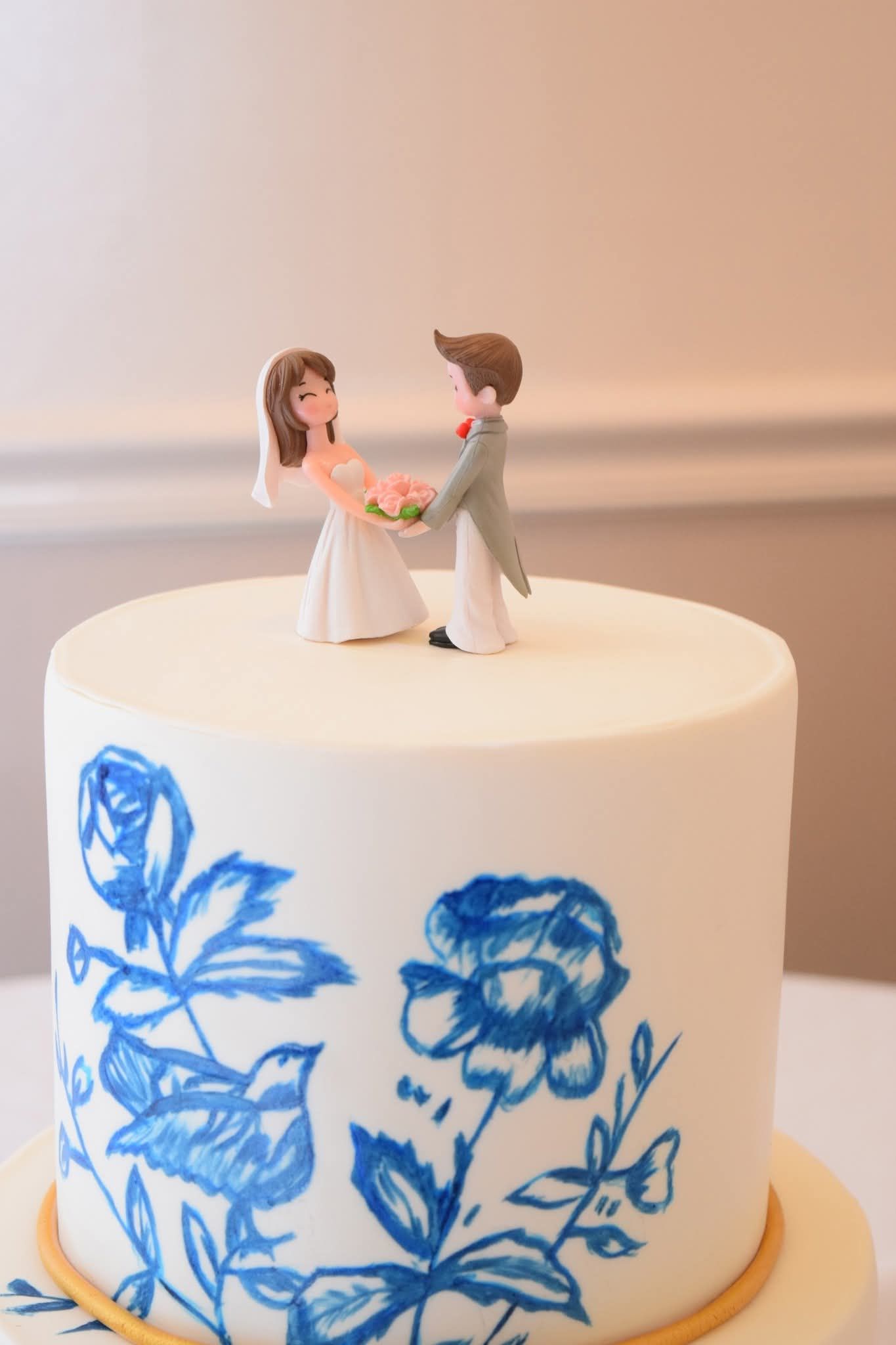 White wedding cake tier topped with bride and groom figurines and blue hand-painted floral designs.
