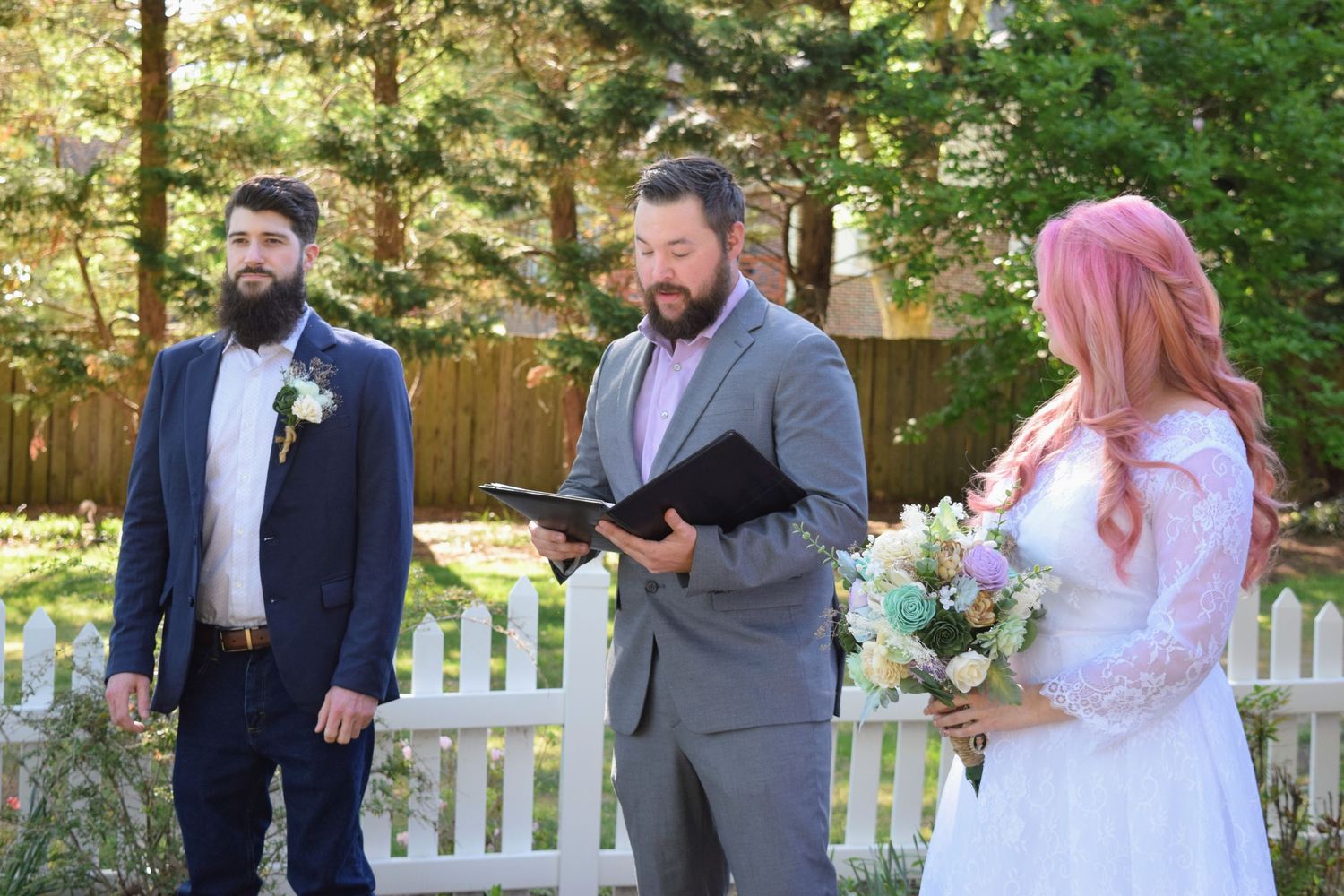 Groom and pink-haired bride stand before officiant during outdoor backyard wedding ceremony near white picket fence.