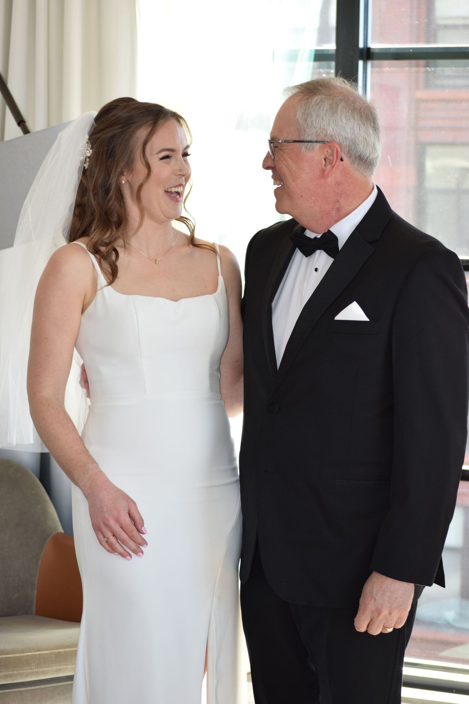 Bride in white gown laughing with older man in tuxedo at wedding venue.