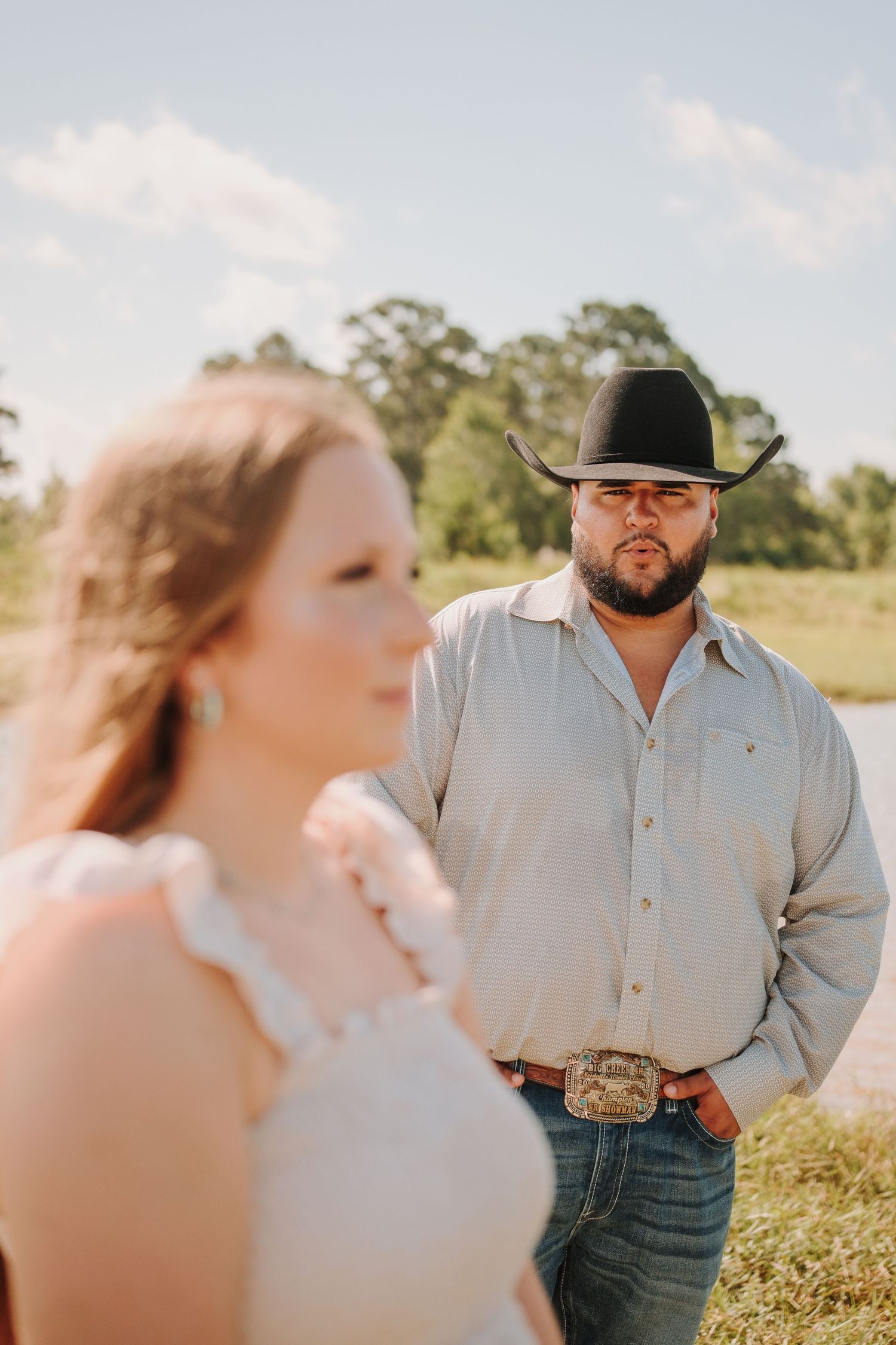 Bearded man in black cowboy hat and jeans stands behind woman in sunny rural outdoor setting.
