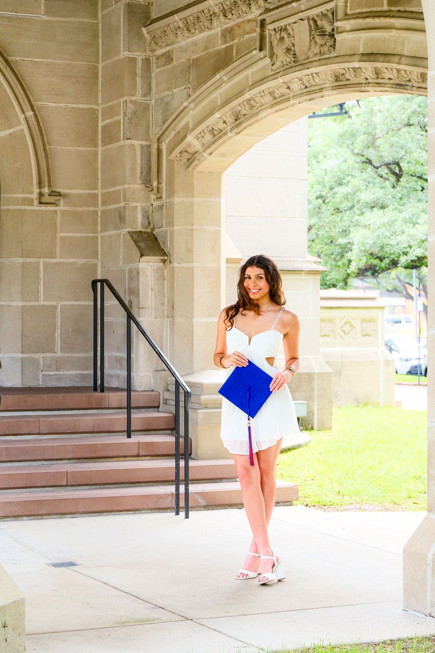 Smiling graduate in white dress holding blue cap under stone archway on college campus.