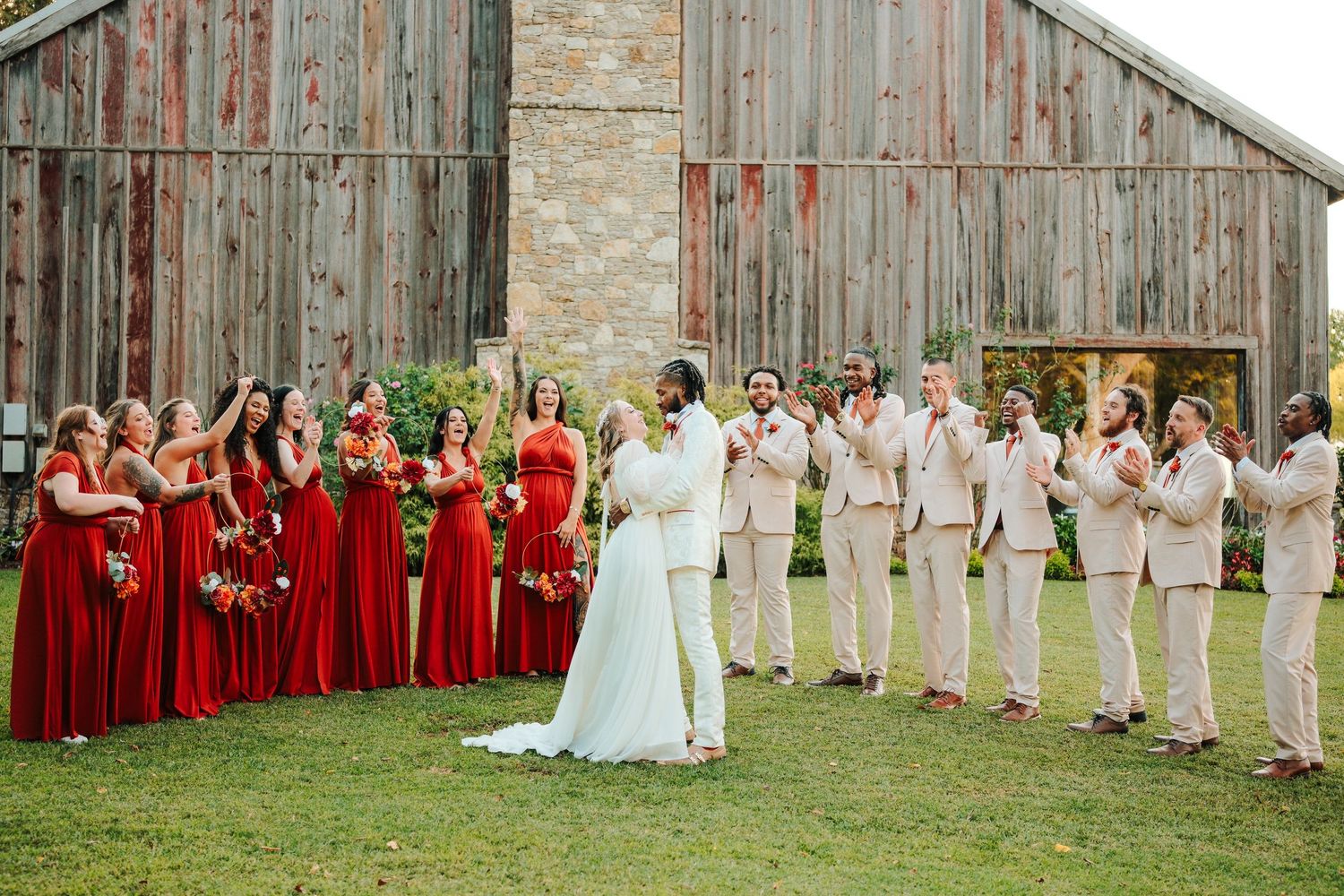 Wedding party in red bridesmaid dresses and tan suits cheering for bride outside rustic barn venue.