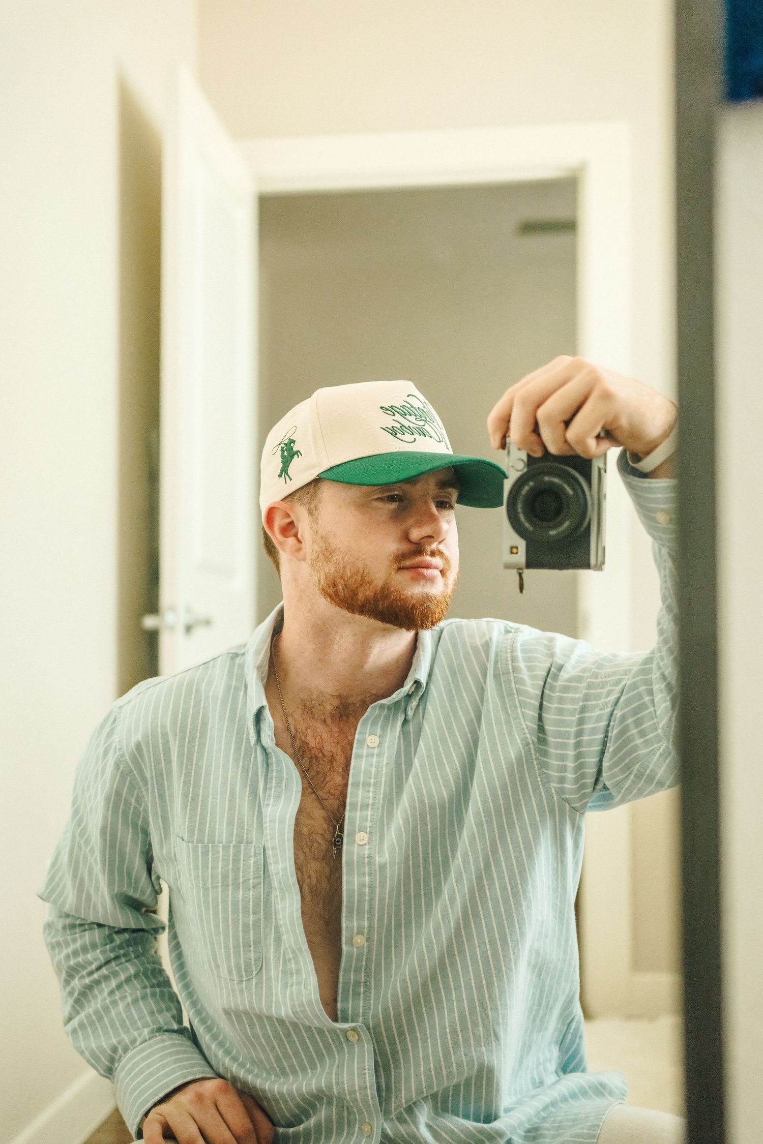 Man in green trucker hat takes mirror selfie with vintage camera, wearing open light blue striped shirt.