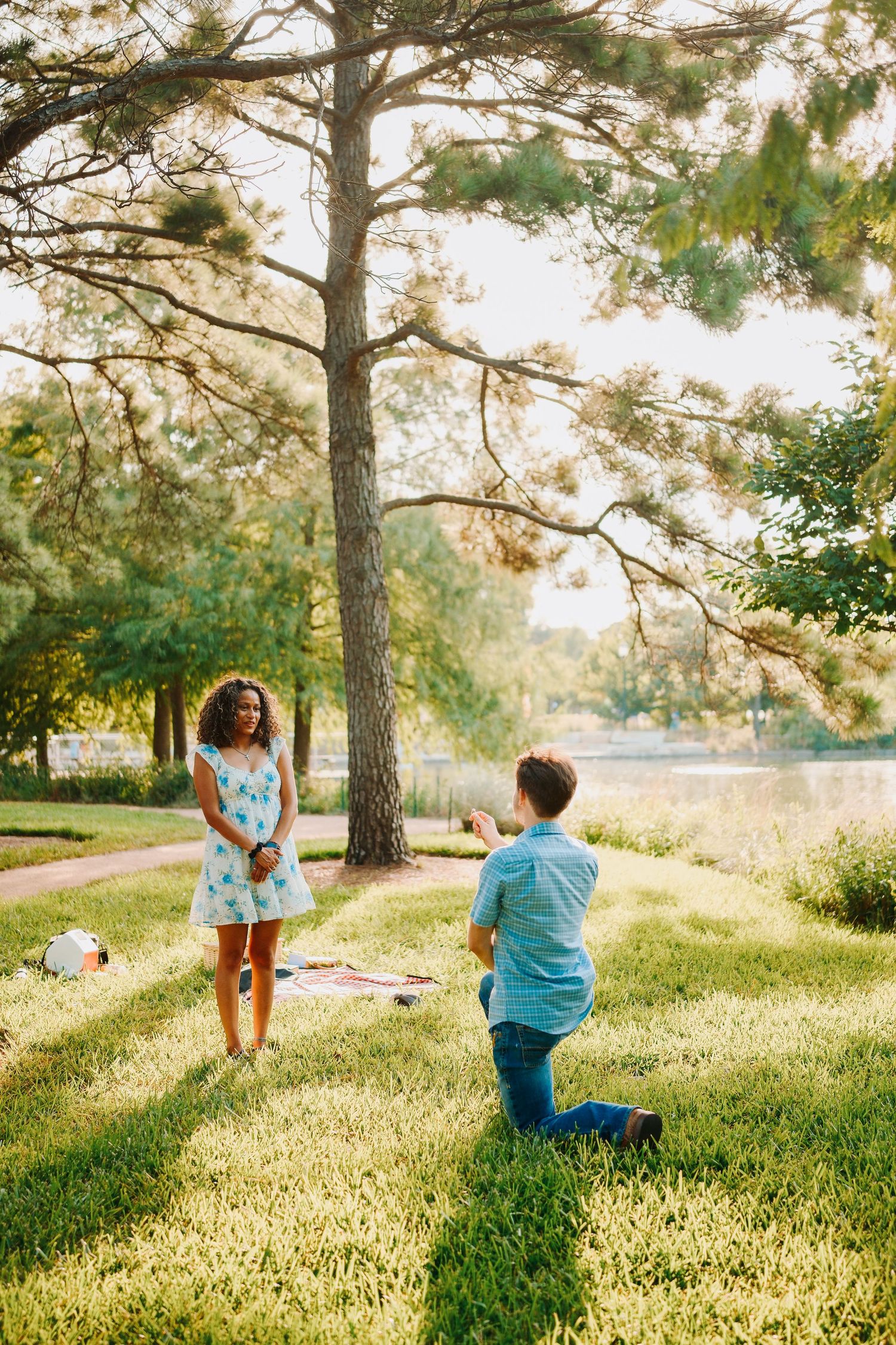 Man kneeling on one knee proposing to woman in blue floral dress in a sunny park by a lake.
