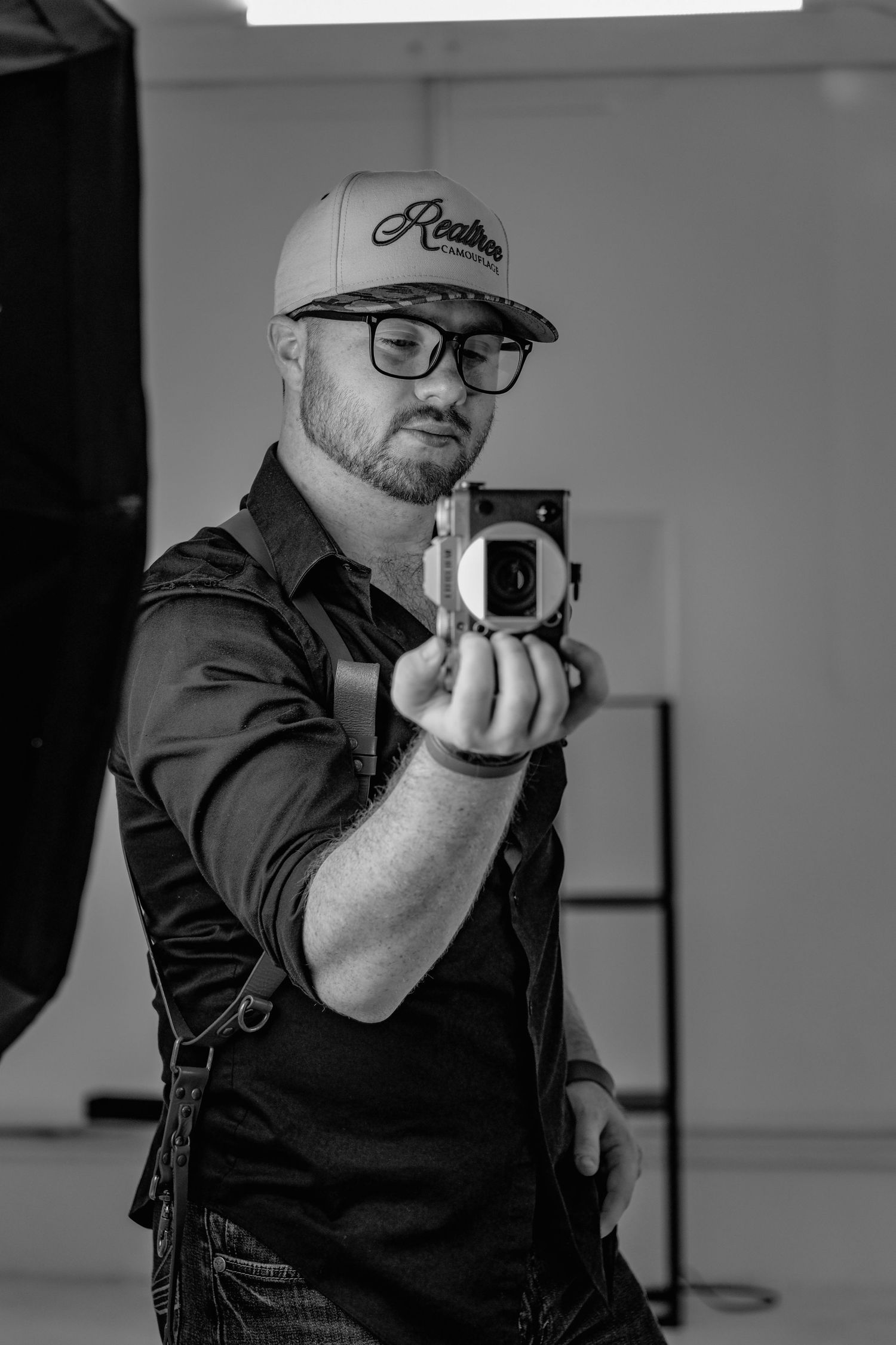 Black and white photo of a bearded man in a cap and glasses taking a mirror selfie with a vintage camera.