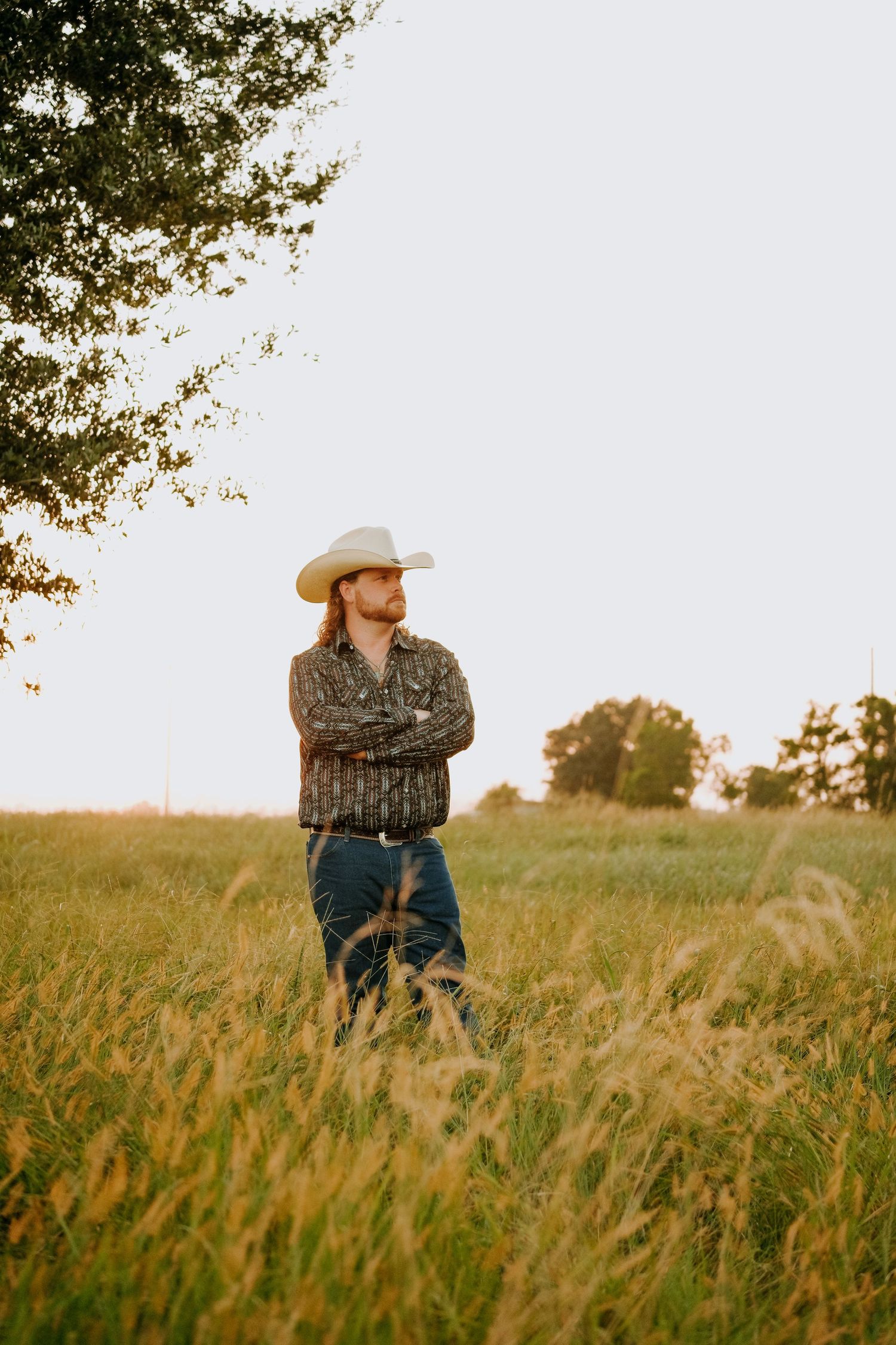 Cowboy in plaid shirt and hat standing in golden grass field at sunset.