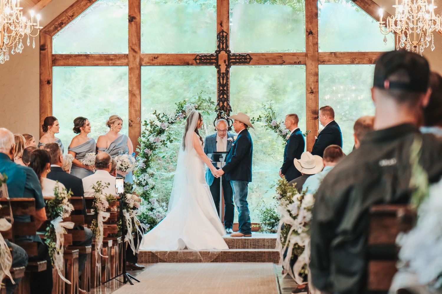 Elegant indoor wedding ceremony with bride and groom at floral altar beneath wooden cross in rustic chapel.