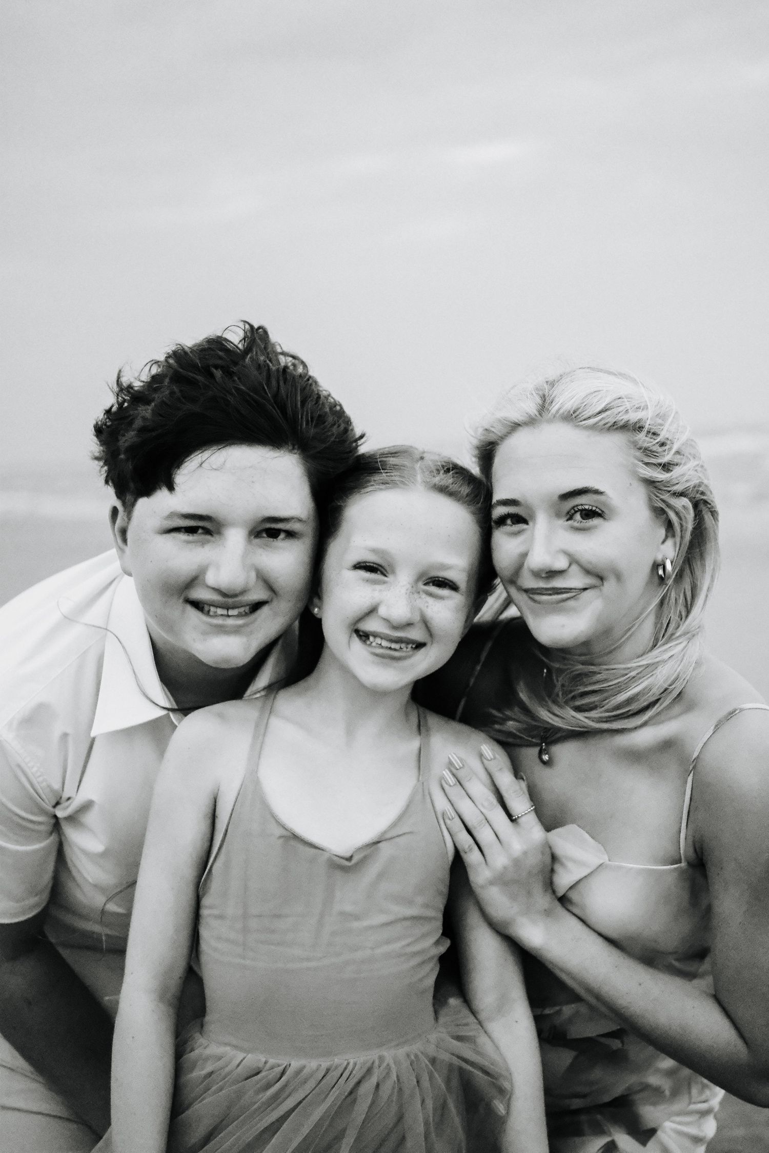Black and white photo of three smiling siblings posing together outdoors near a beach.