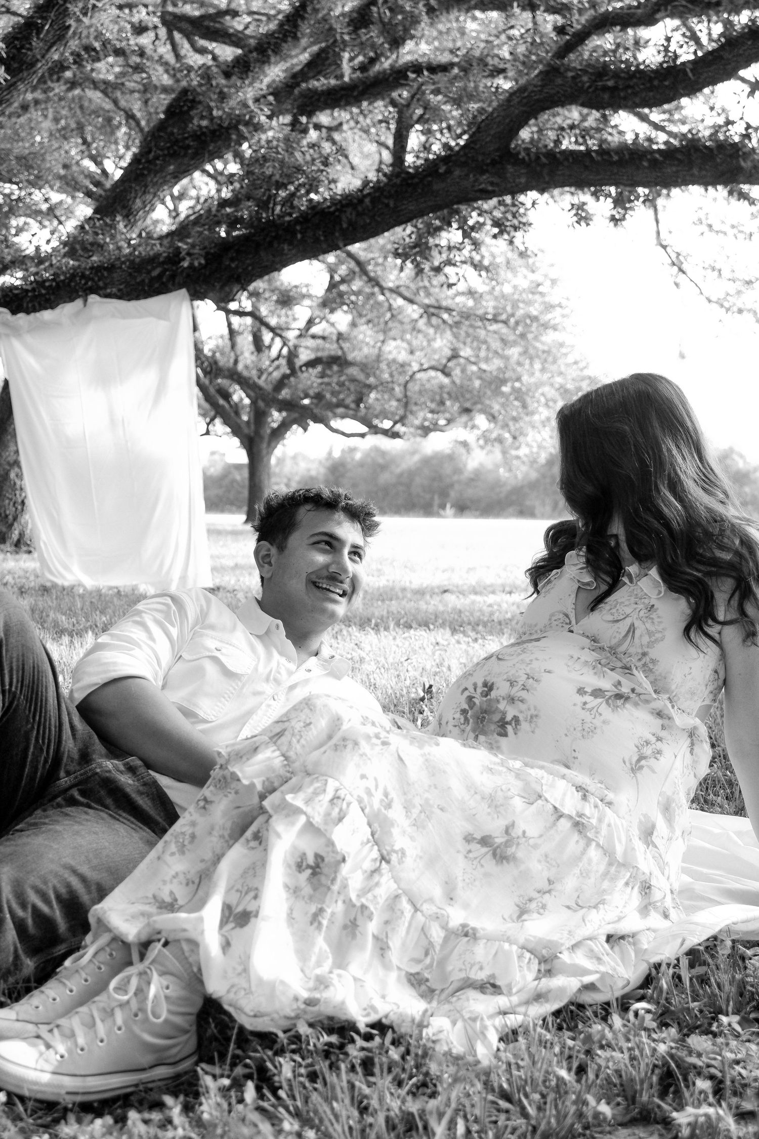 Black and white photo of smiling couple relaxing on grass under a large oak tree outdoors.