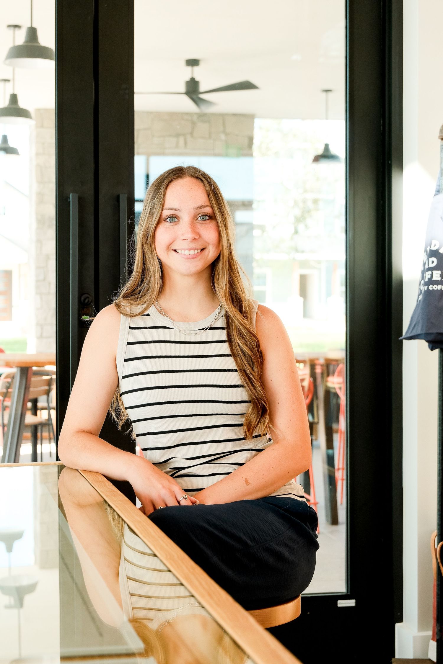 Smiling young woman in striped top leaning on wooden railing inside modern coffee shop.