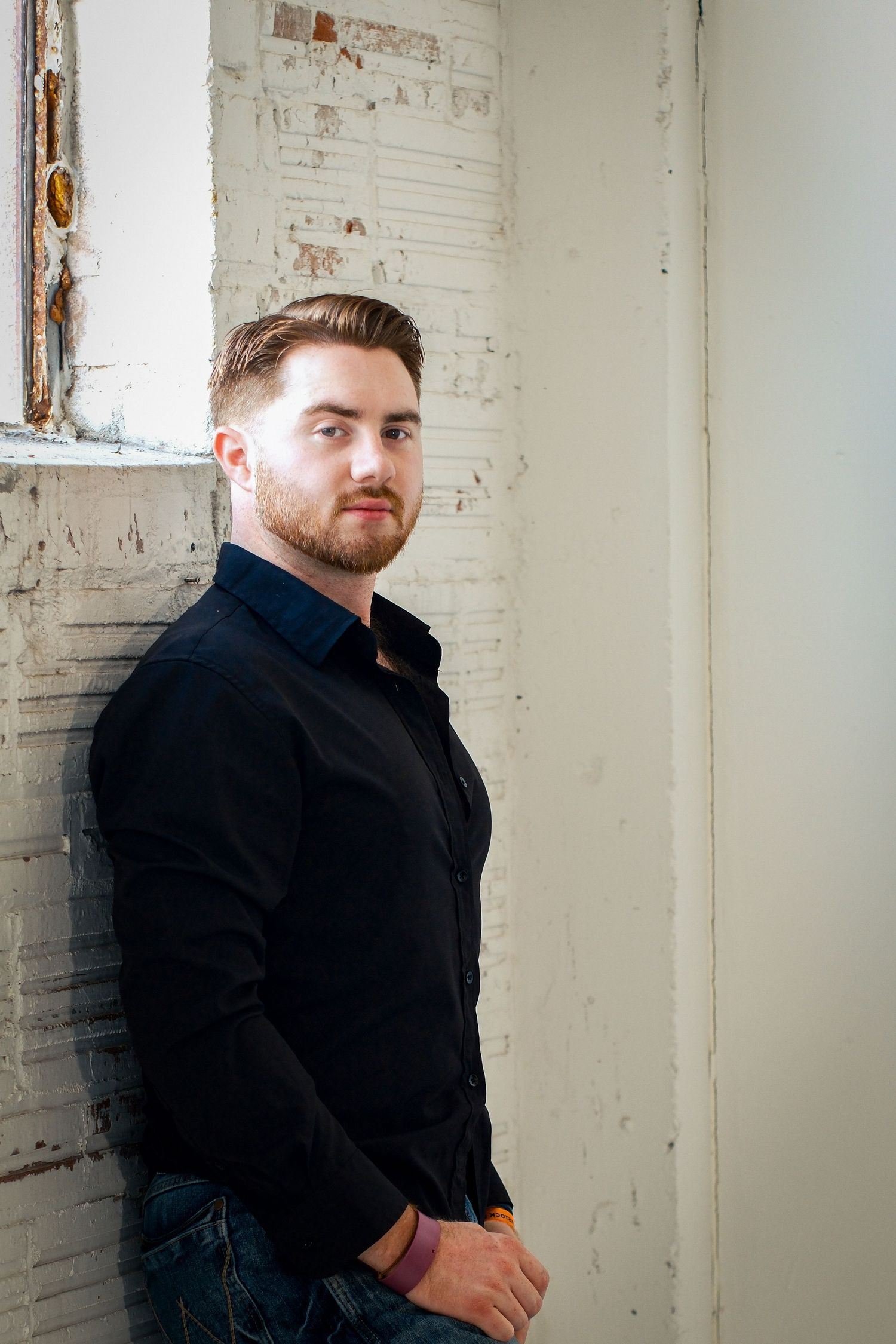 Confident young bearded man in black shirt posing against a weathered white brick wall.