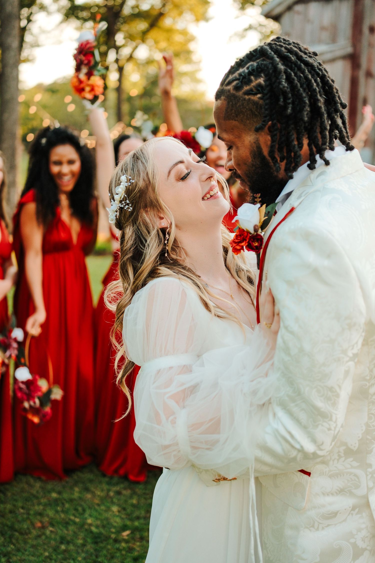A bride and groom share a romantic moment outdoors, surrounded by bridesmaids in red dresses at their wedding.