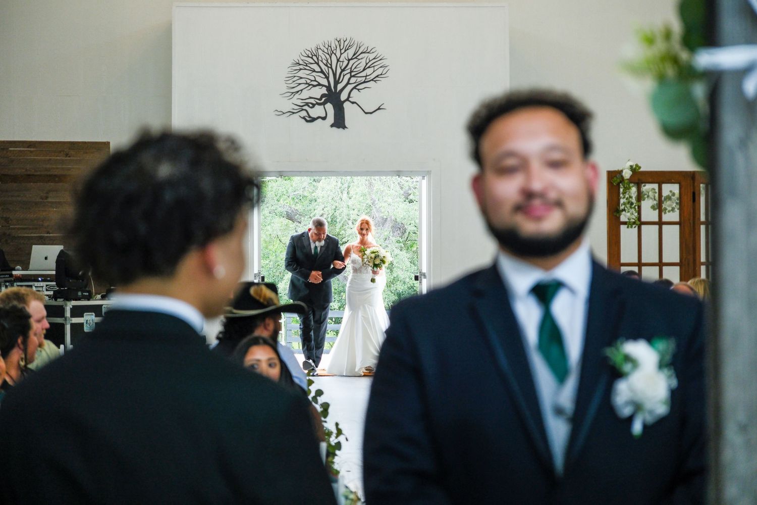 Groom smiling in foreground as bride walks down aisle with father in bright white wedding venue.