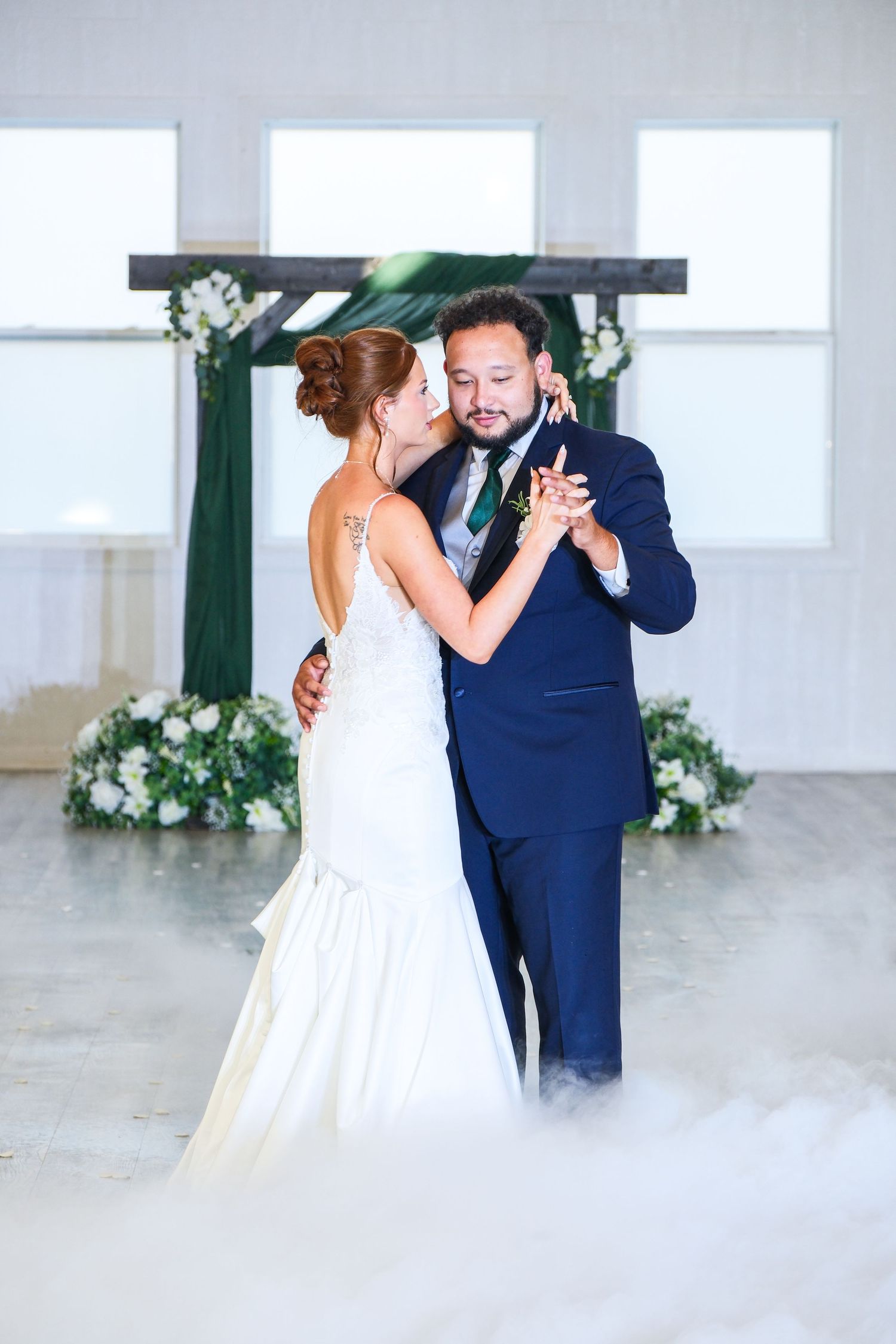 Bride and groom share first dance surrounded by romantic fog effects beneath floral cross backdrop.