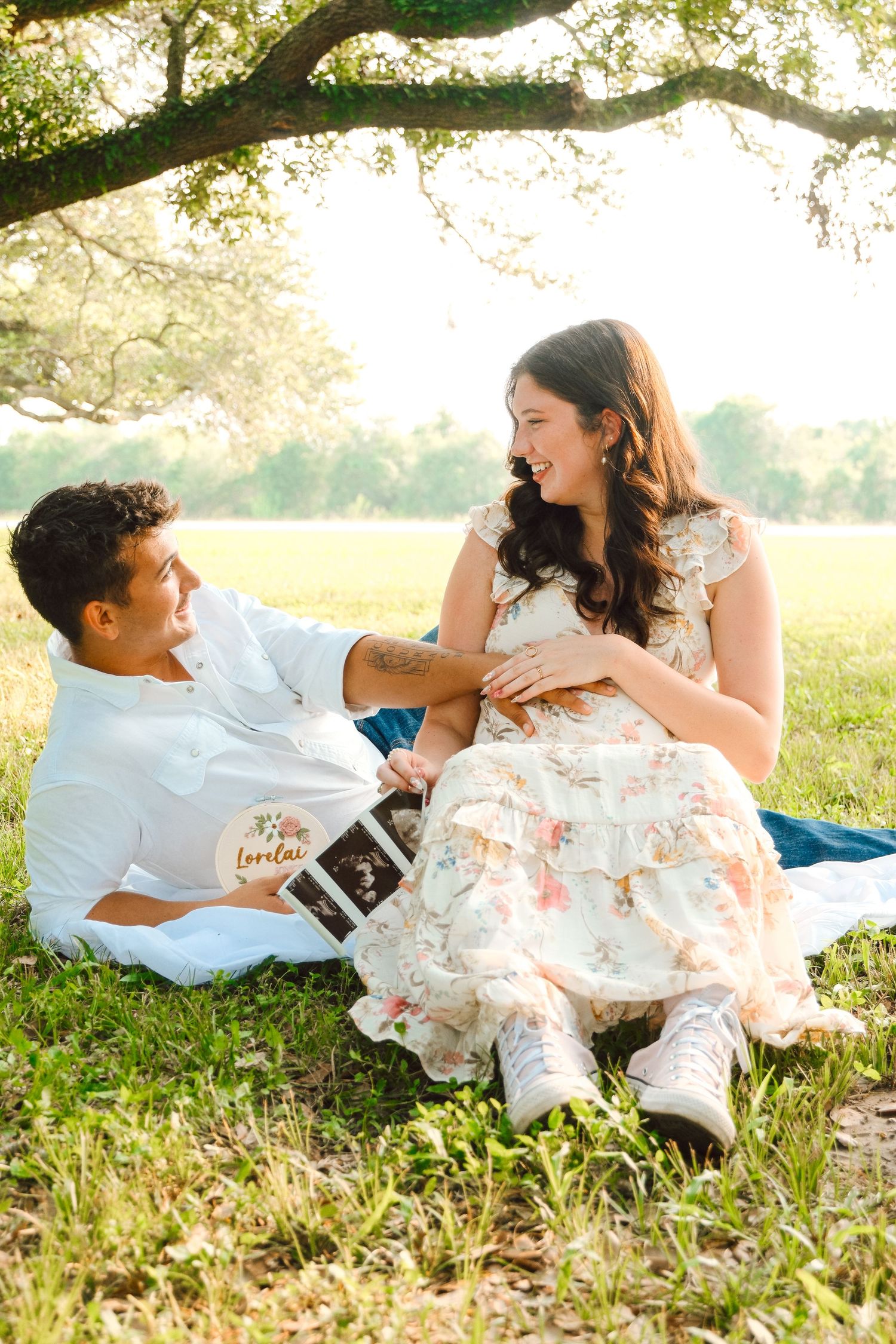 Couple sitting under oak tree sharing pregnancy ultrasound photo in sunny outdoor setting.