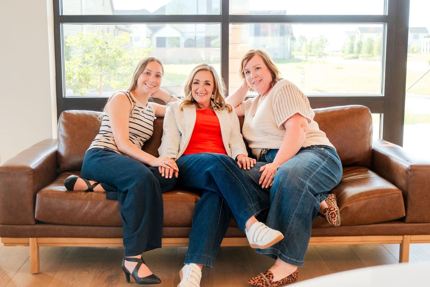 Three women smiling together on a brown leather couch in a bright modern room.