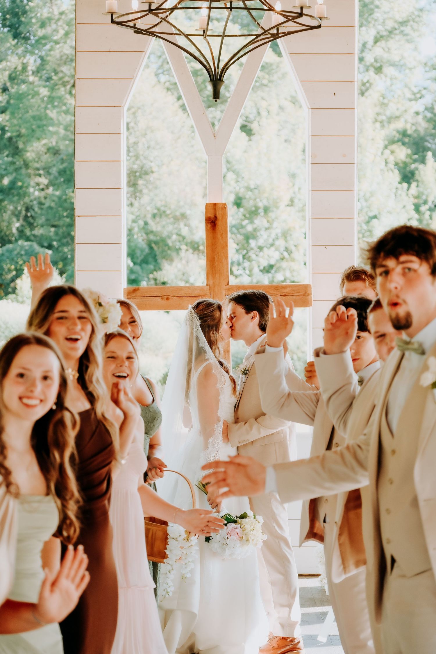 A joyful wedding party celebrates as the bride and groom share their first kiss beneath a wooden cross in a sunlit chapel.