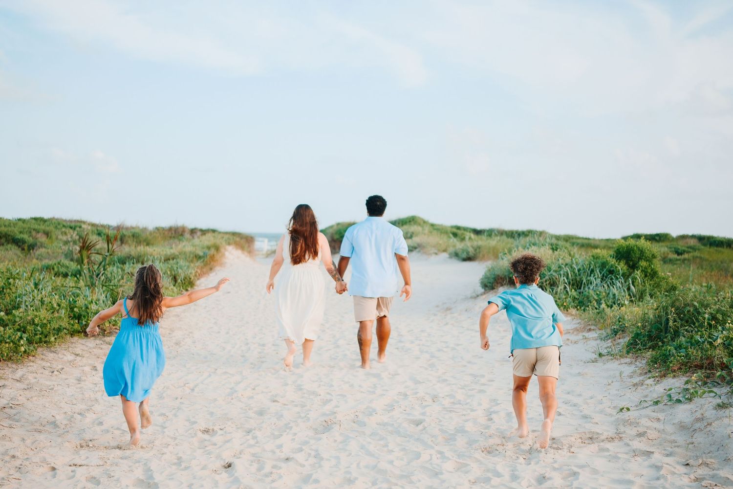 Family of four walking hand-in-hand toward a sandy beach path surrounded by green dunes under a blue sky.