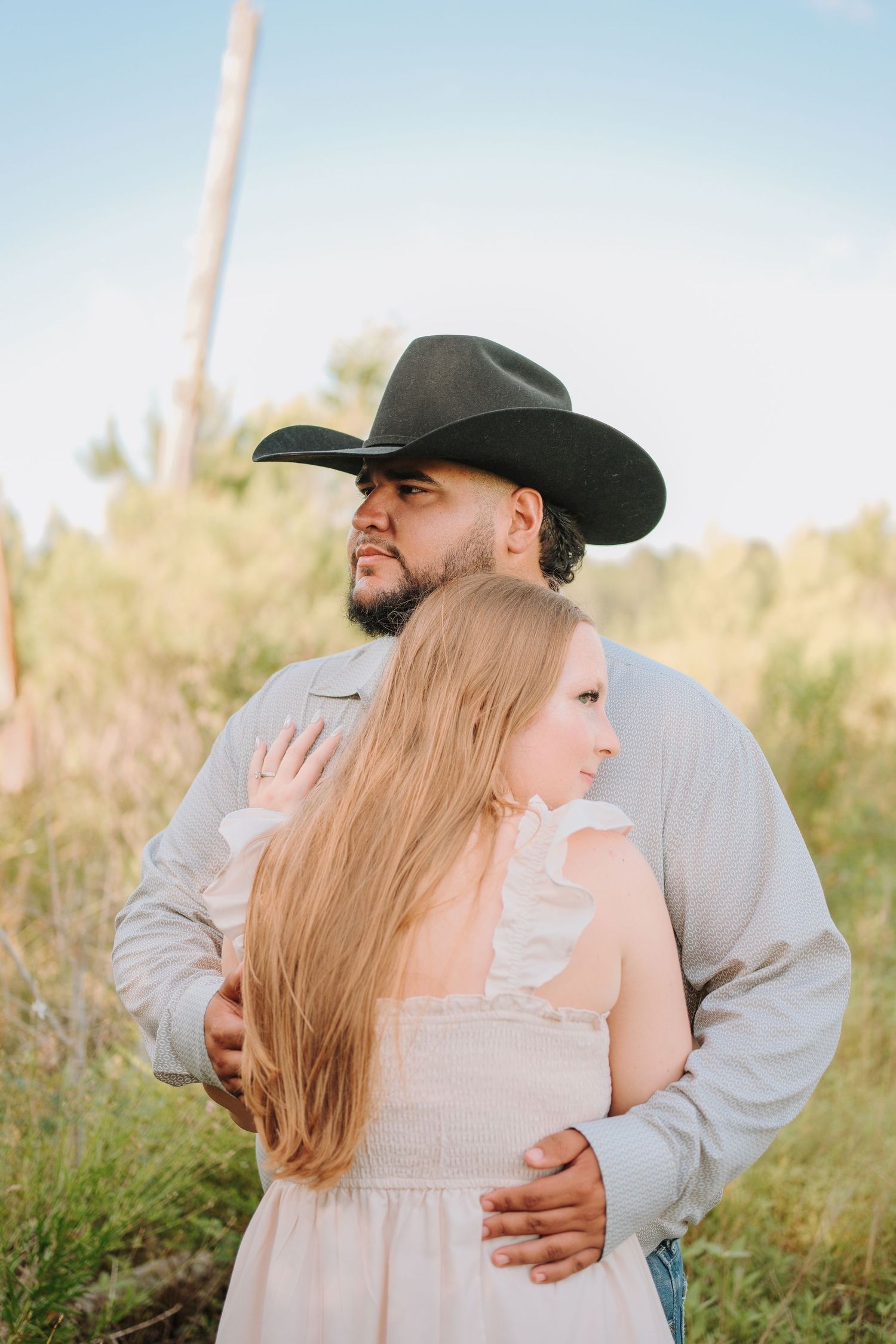 A couple embraces outdoors, man wearing black cowboy hat and gray shirt, woman in pink dress with long blonde hair.
