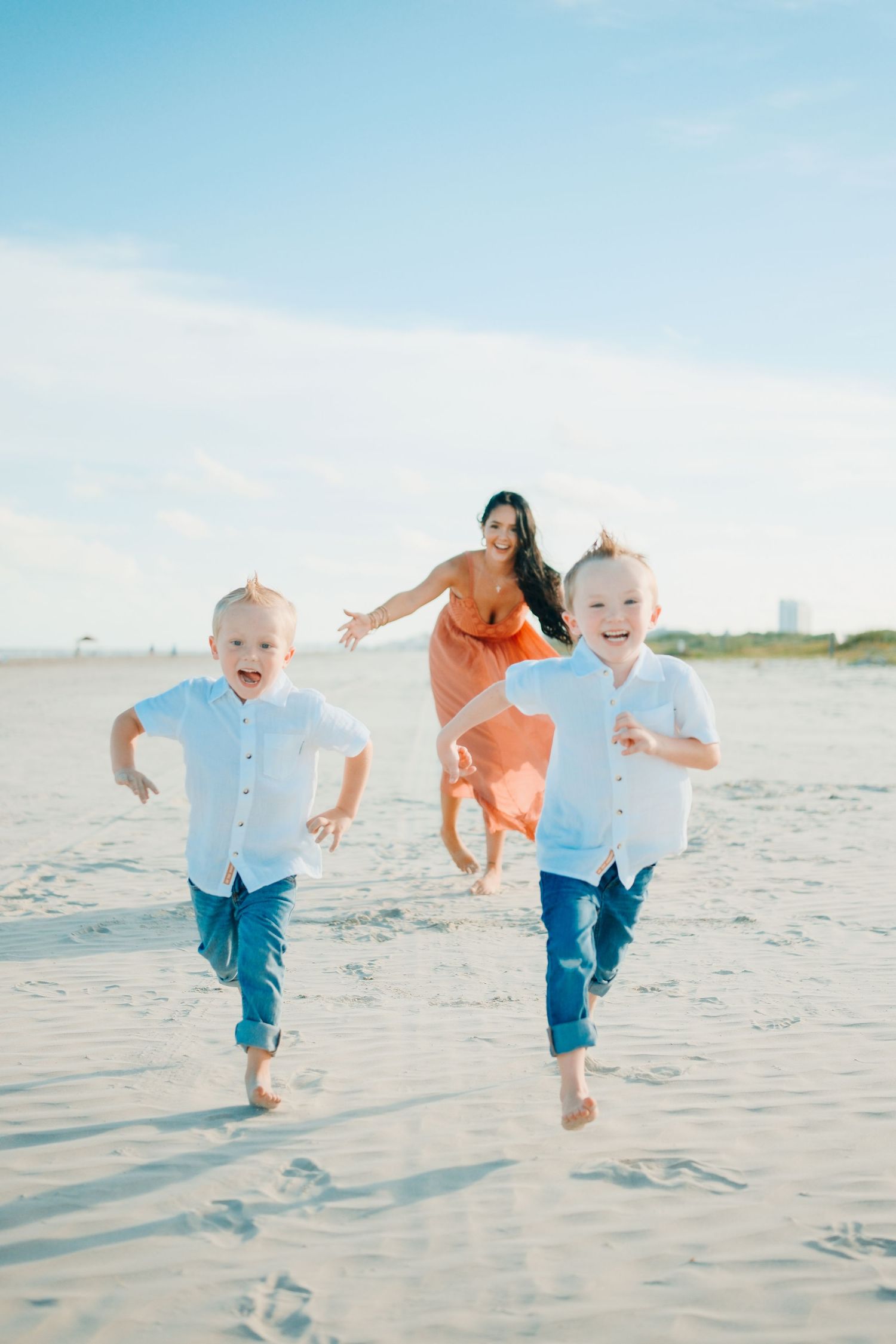 A joyful mother in an orange dress chases two laughing boys in white shirts on a sunny beach.