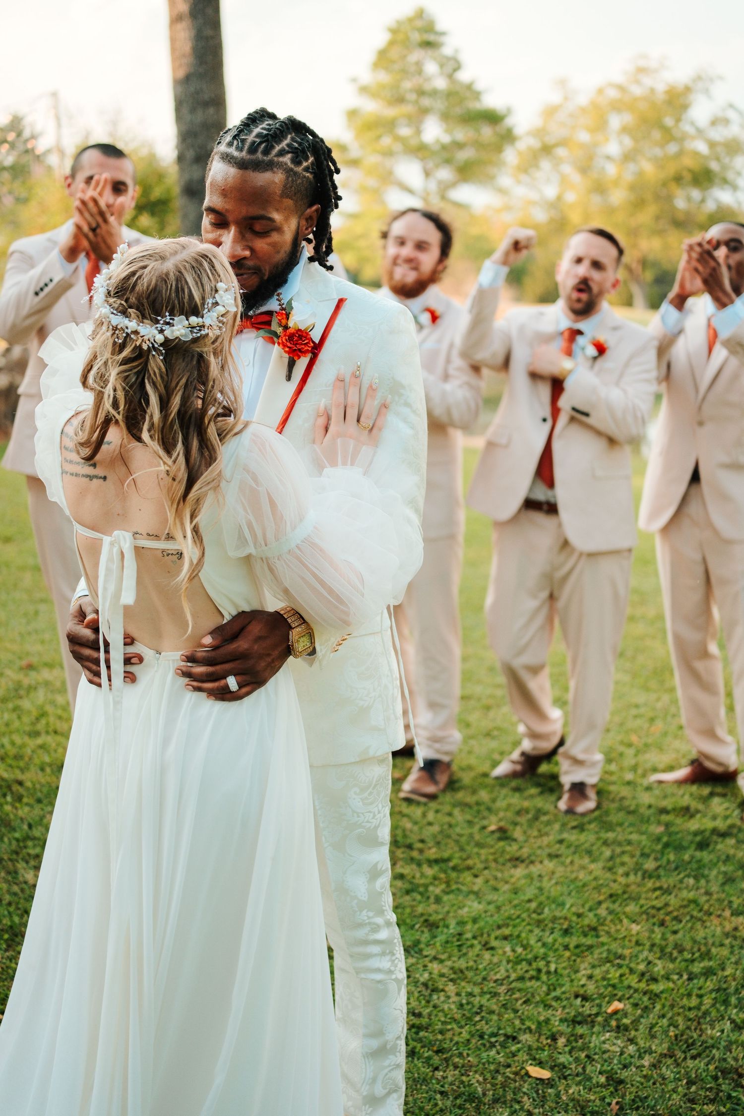 Bride and groom share first kiss at outdoor wedding ceremony while groomsmen in tan suits celebrate behind them.