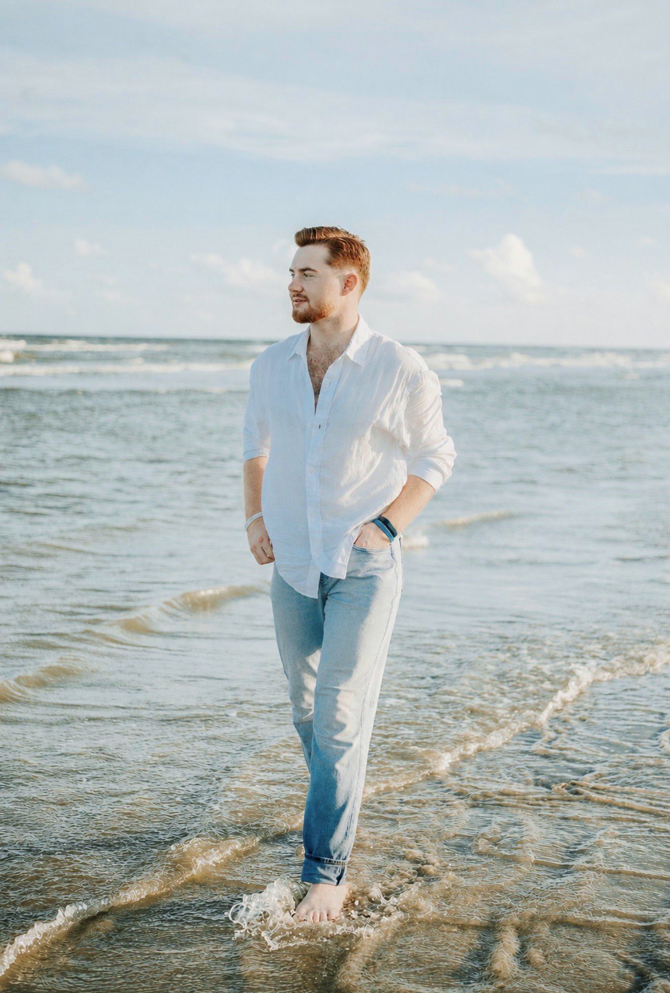 Young man in white linen shirt and jeans walking barefoot in ocean waves on a sunny beach.
