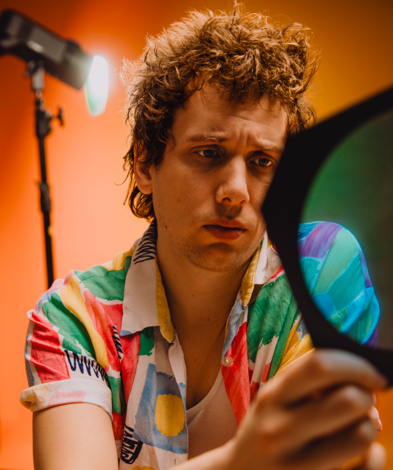 Close-up of curly-haired young man in vibrant patterned shirt looking into mirror in orange studio.