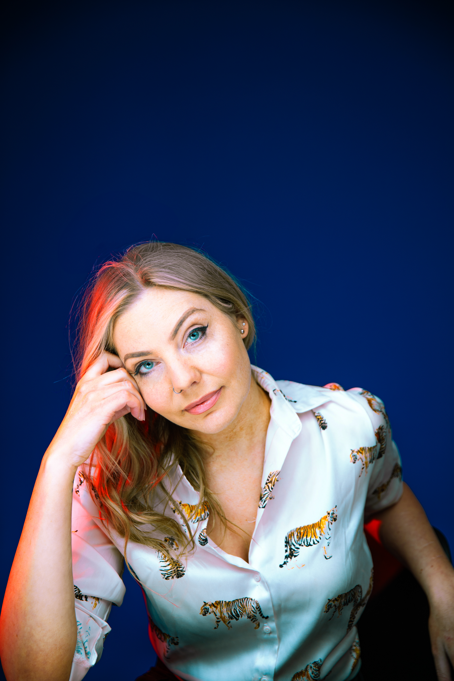 Blonde woman with blue eyes in tiger-print shirt posing against deep navy blue studio background.