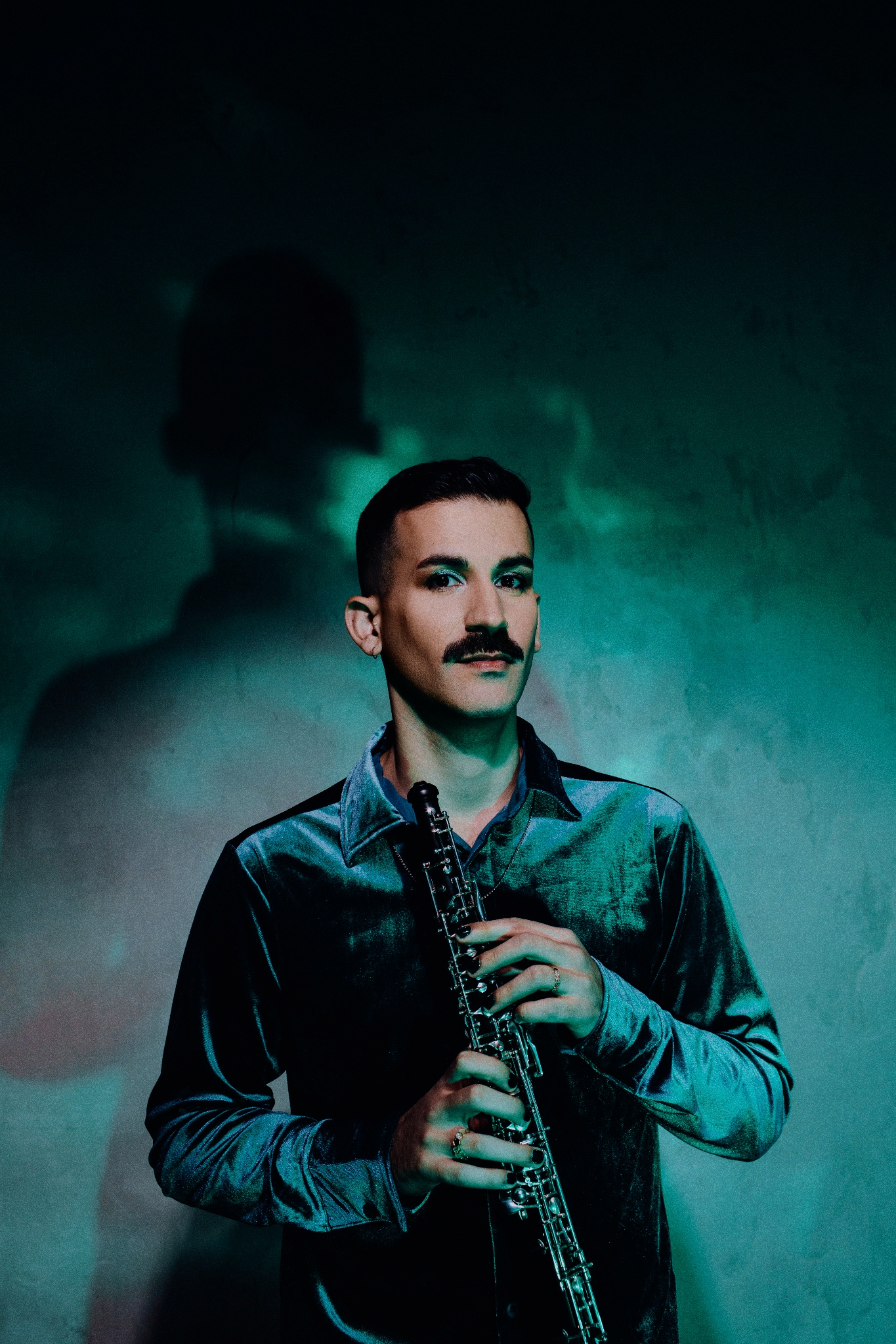 Mustachioed male musician holding oboe against teal smoky wall with dramatic shadow behind him.